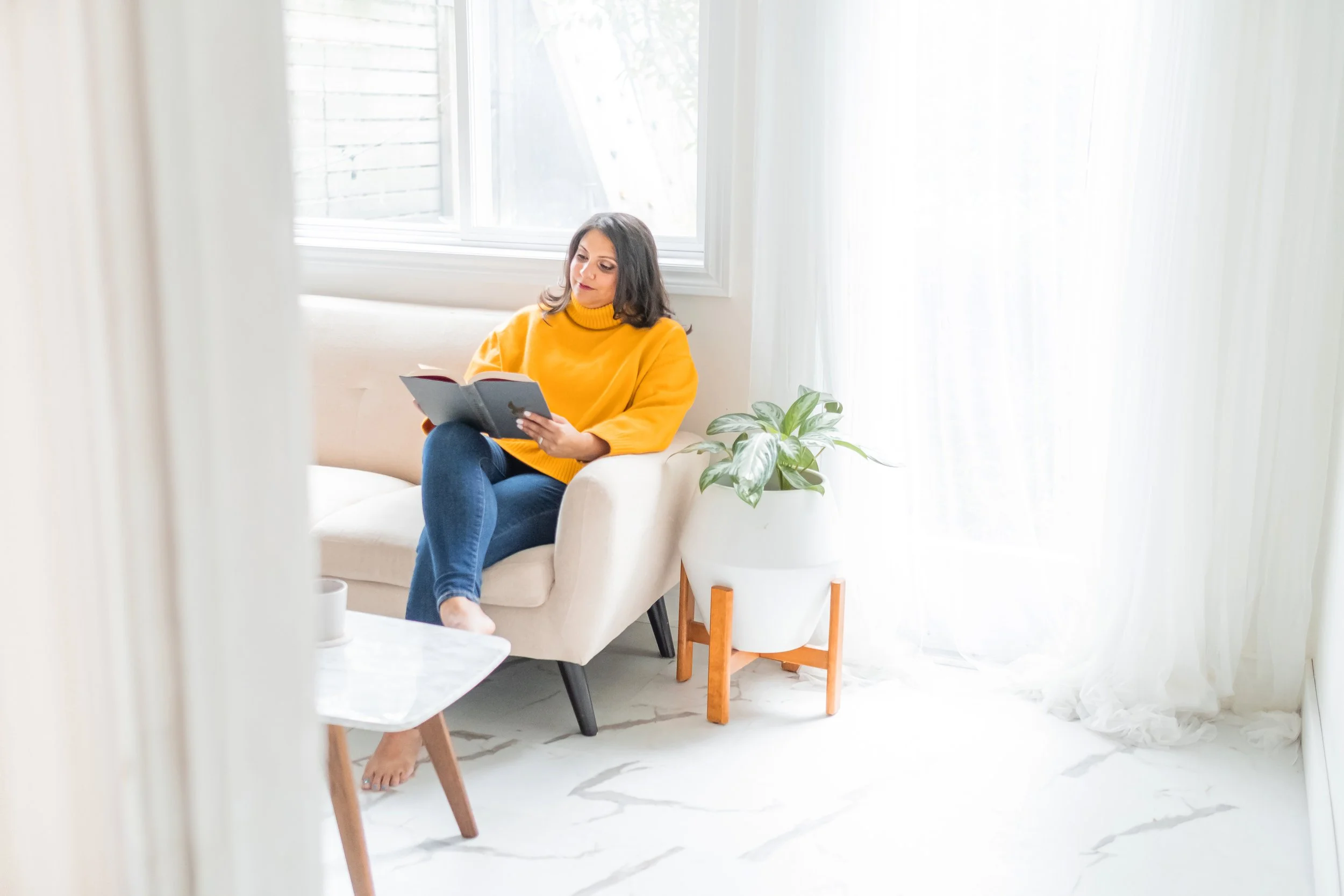 Bright image of Kiren sitting on a white couch reading a book. Wearing a bright yellow sweater and blue jeans.