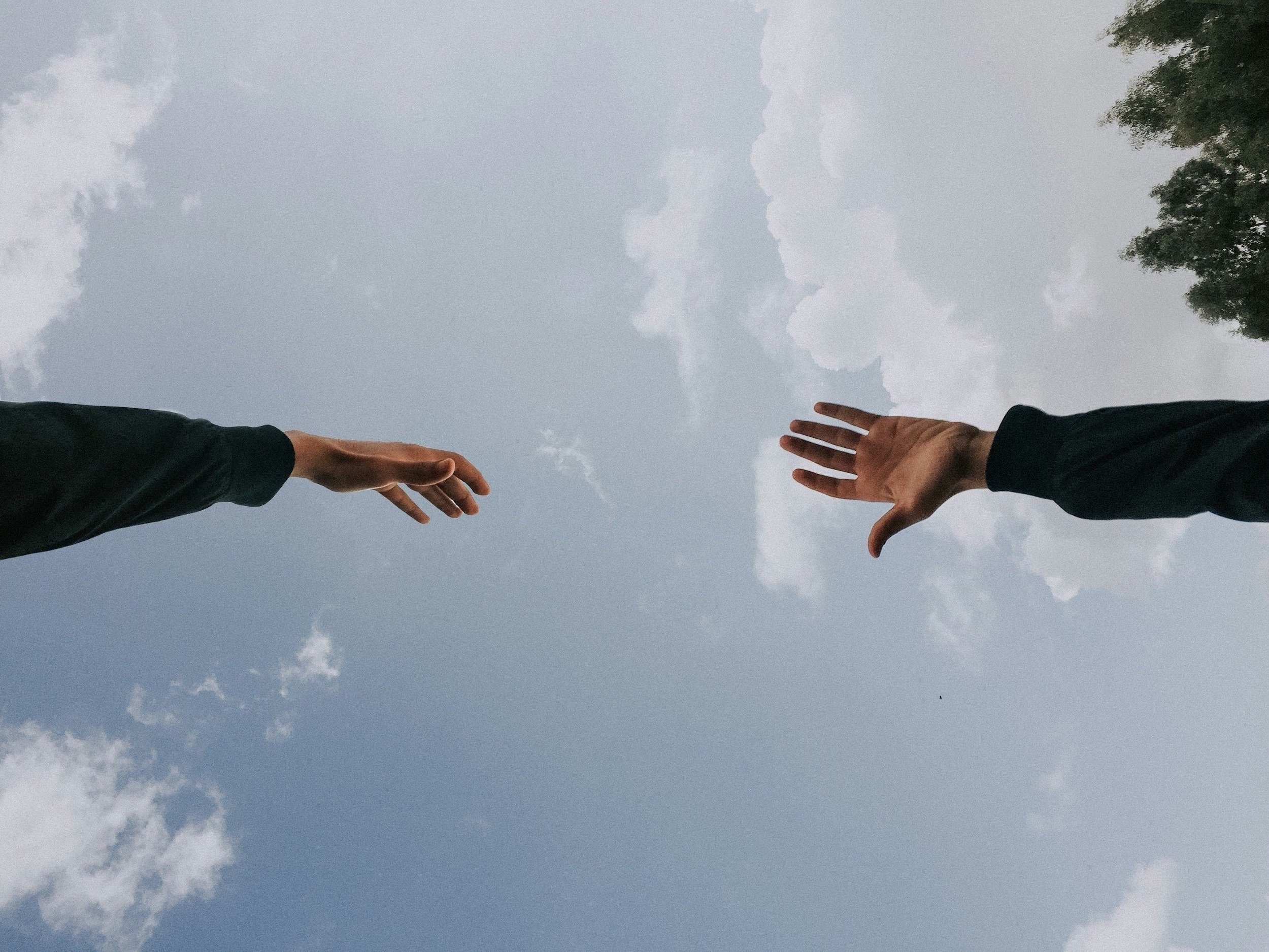 Image of two hands reaching towards each other. The sky in the background is blue with some clouds.