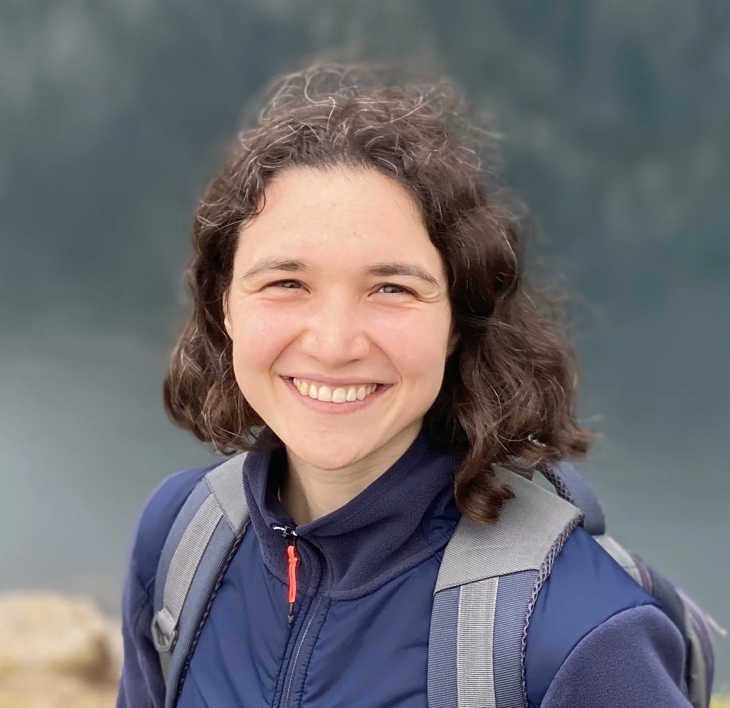 Smiling woman with curly hair wearing a blue jacket and backpack outdoors.