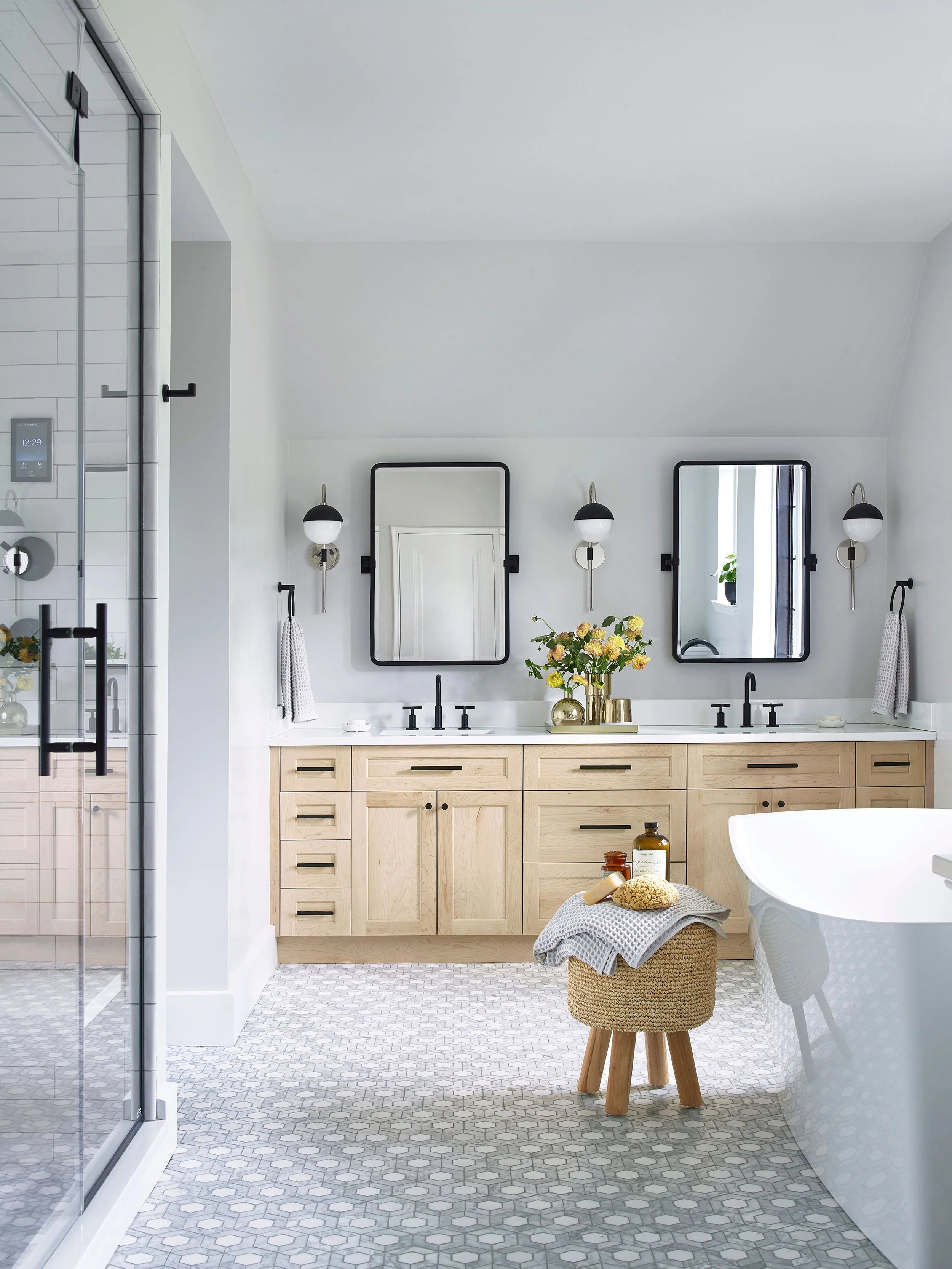 Modern bathroom with light wood vanity, two black-framed mirrors, black wall sconces, a corner with a freestanding tub, and a woven stool with toiletries. Tiled floor and white walls.