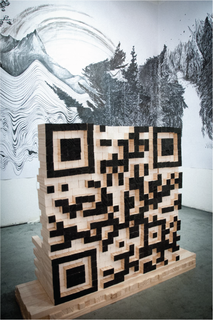 A wooden sculpture of a large QR code made of wood. 70 x 70 x 70 cm. Behind a large format drawing on charcoal, an homage of the German Romantic landscape tradition in crossover with the aesthetics of digital error.