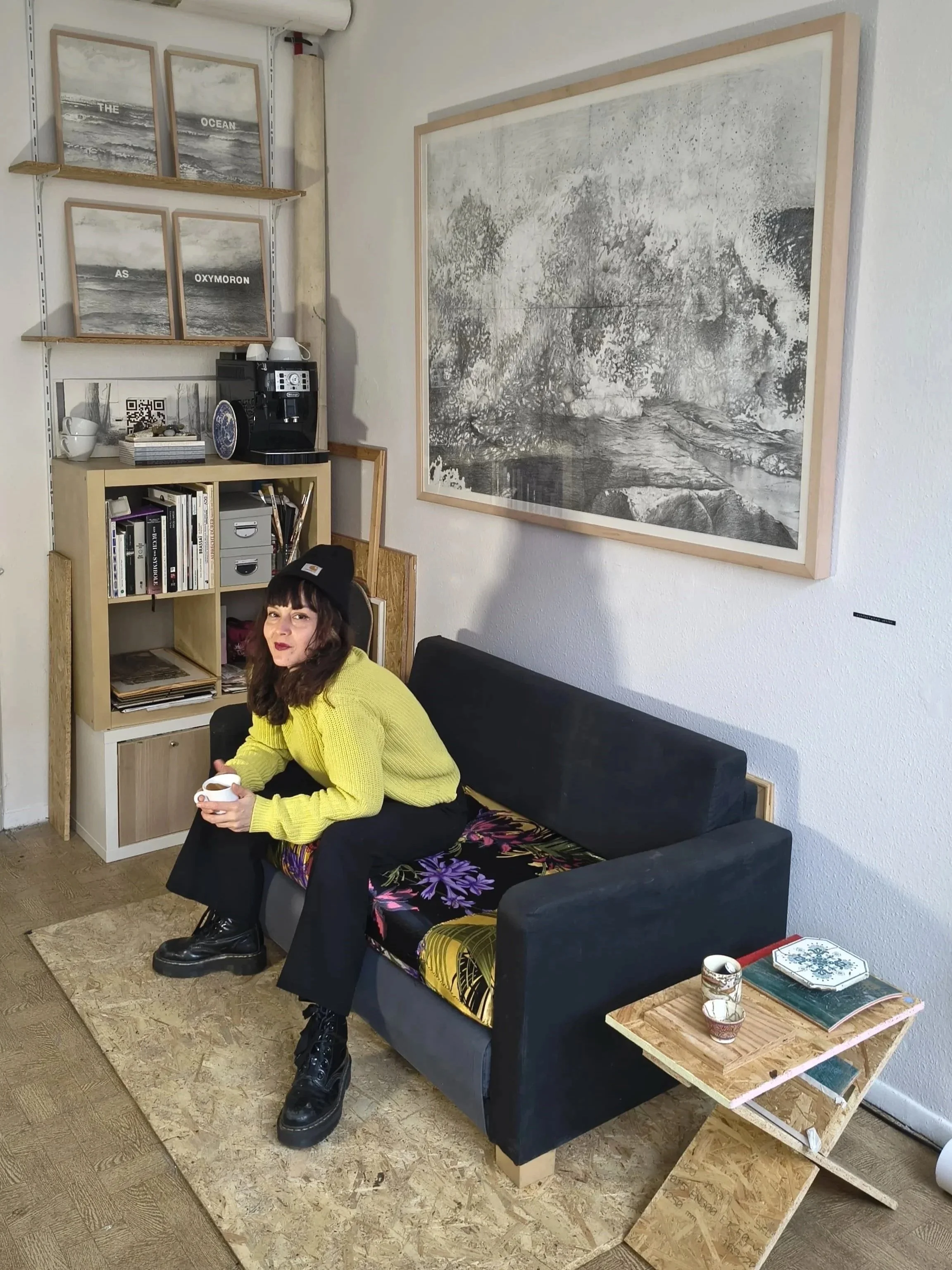 Guillermina Gómez Arévalo, Founder of Studio SATO wearing a yellow sweater, black pants, and black boots, sitting on a black couch in a cozy corner of the studio, on views the series " The Ocean as Oxymoron".