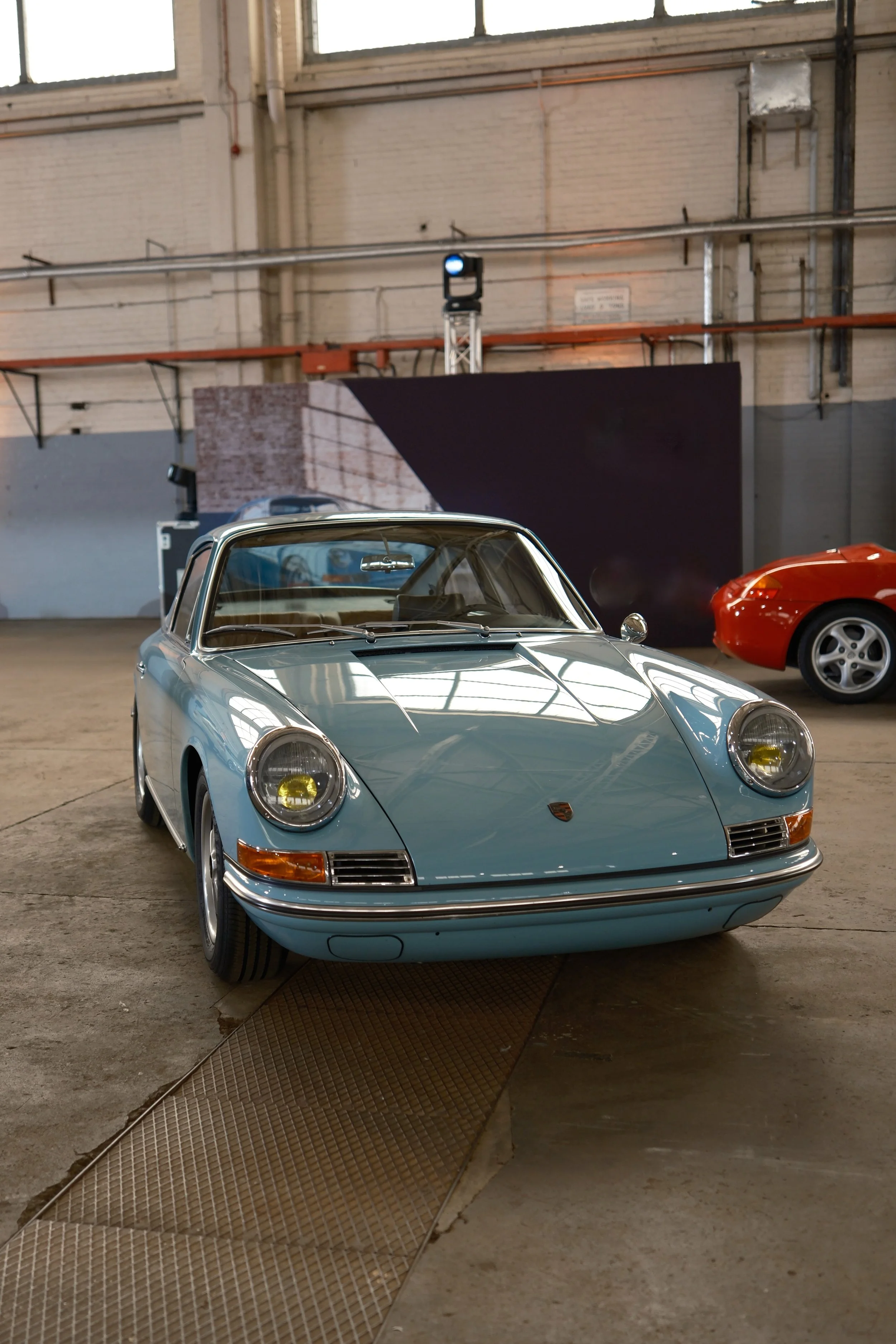 A classic silver Porsche sports car on display indoors, with a red sports car partially visible in the background, in what appears to be a showroom or storage facility with industrial design elements.