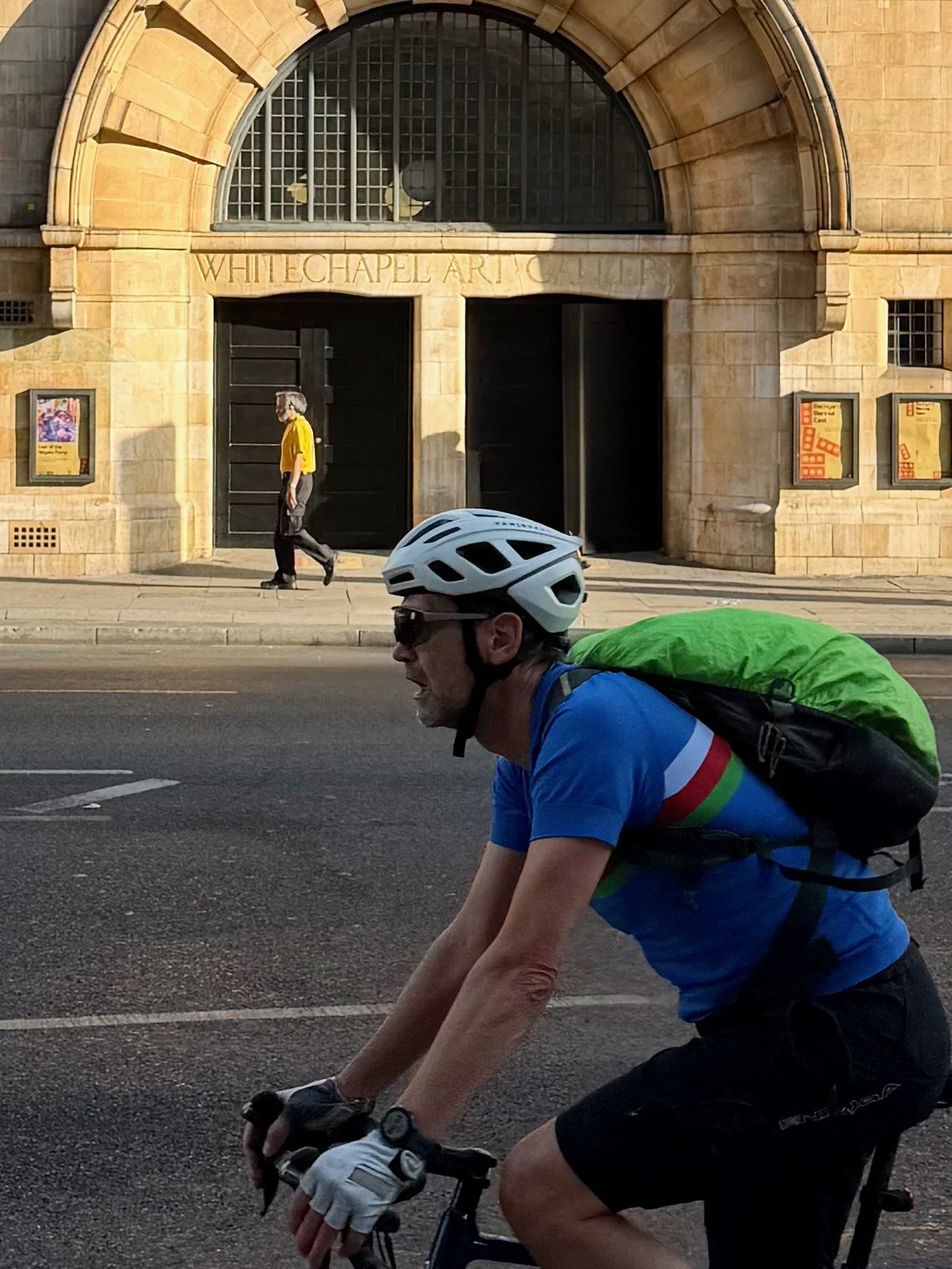 A man riding a bicycle on the street wearing a white helmet, sunglasses, and a blue cycling jersey with rainbow stripes, carrying a green backpack. In the background, a woman in a yellow top walks past the Whitechapel Art Gallery entrance.