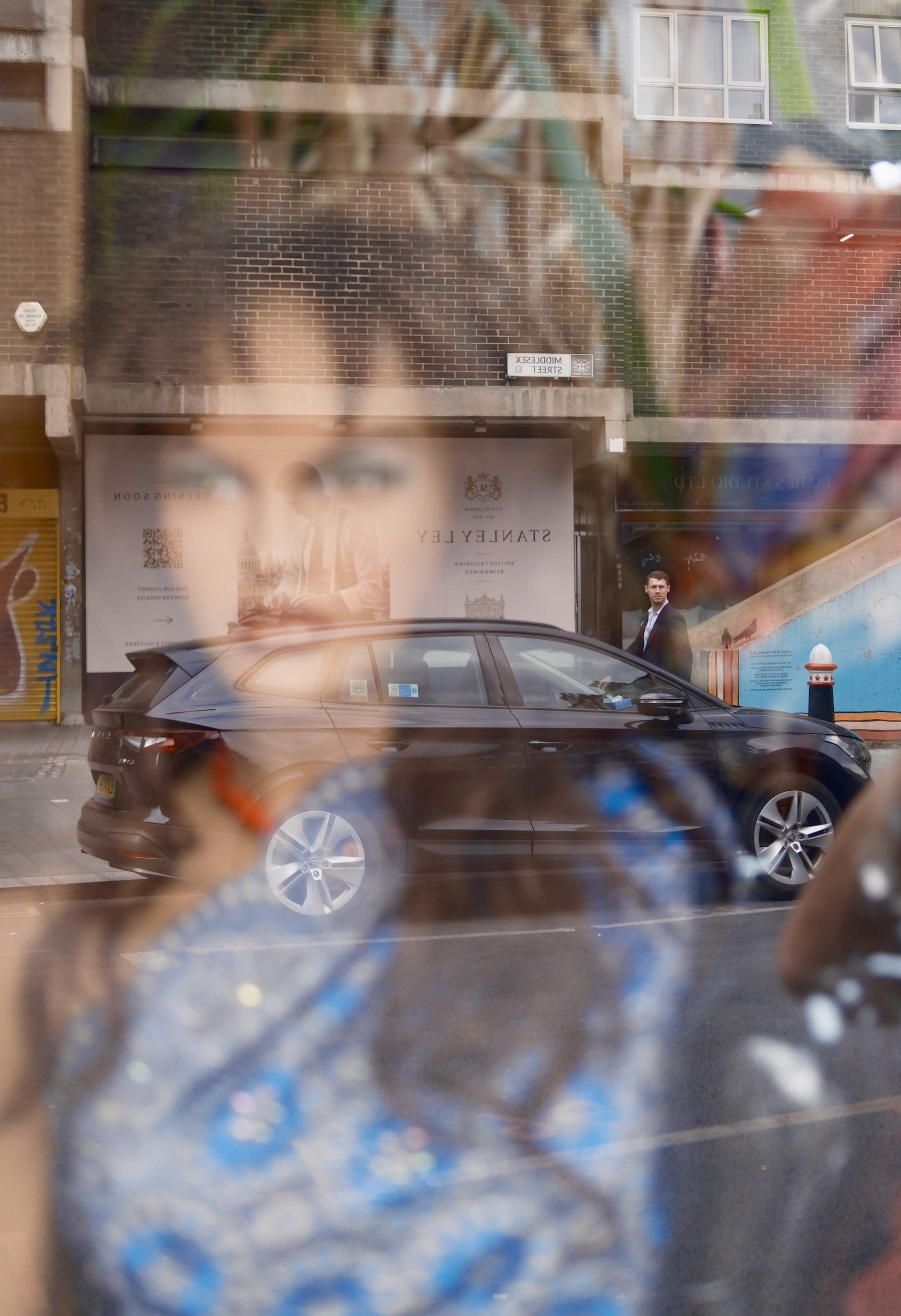 View through a window showing a brick building facade, a parked black car, and a man standing on the sidewalk. The window has reflections and distortions, partially obscuring the scene.