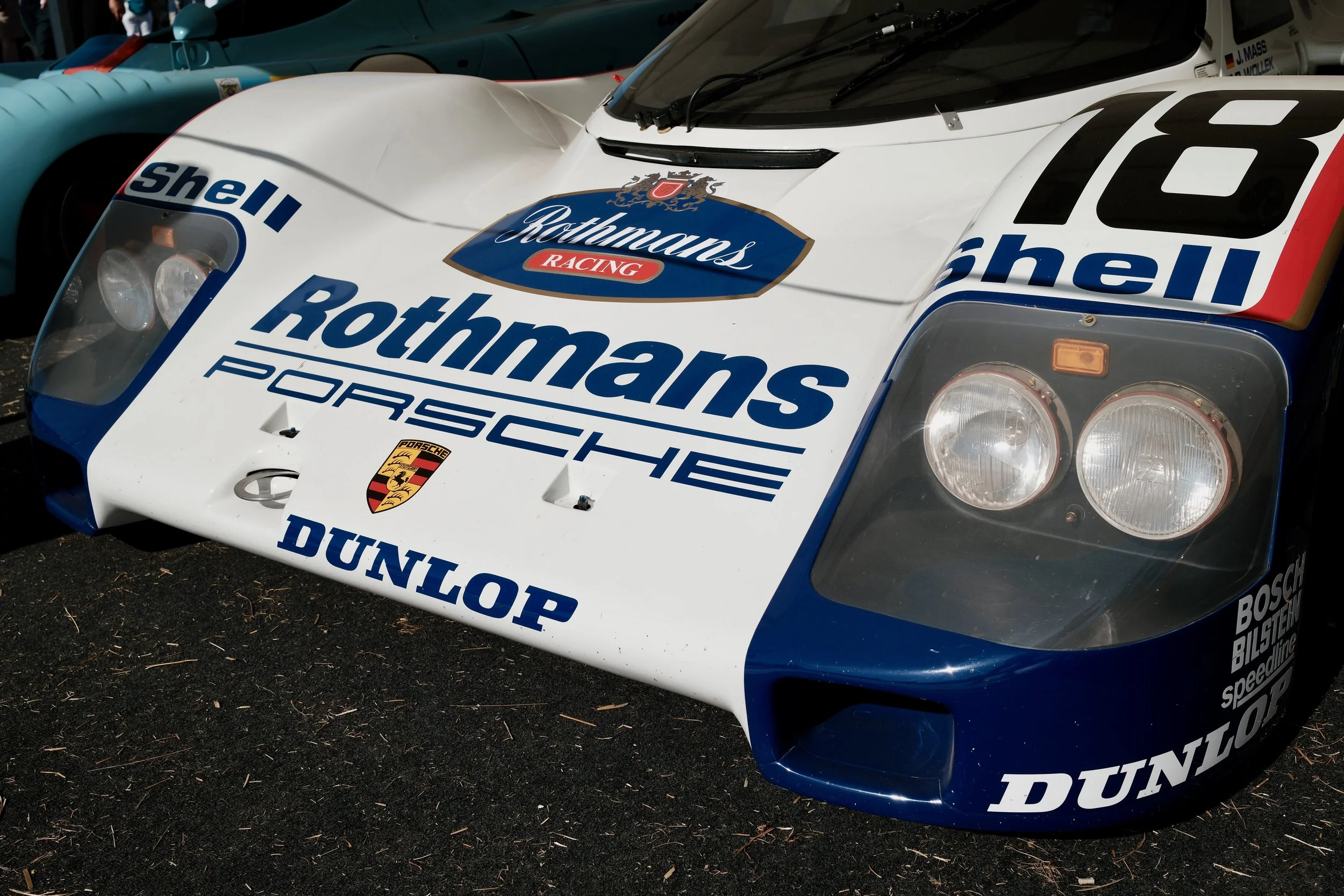 Close-up of a vintage race car with sponsor logos including Rothmans, Shell, and Porsche, featuring a white, blue, and red color scheme, parked on a dirt surface.