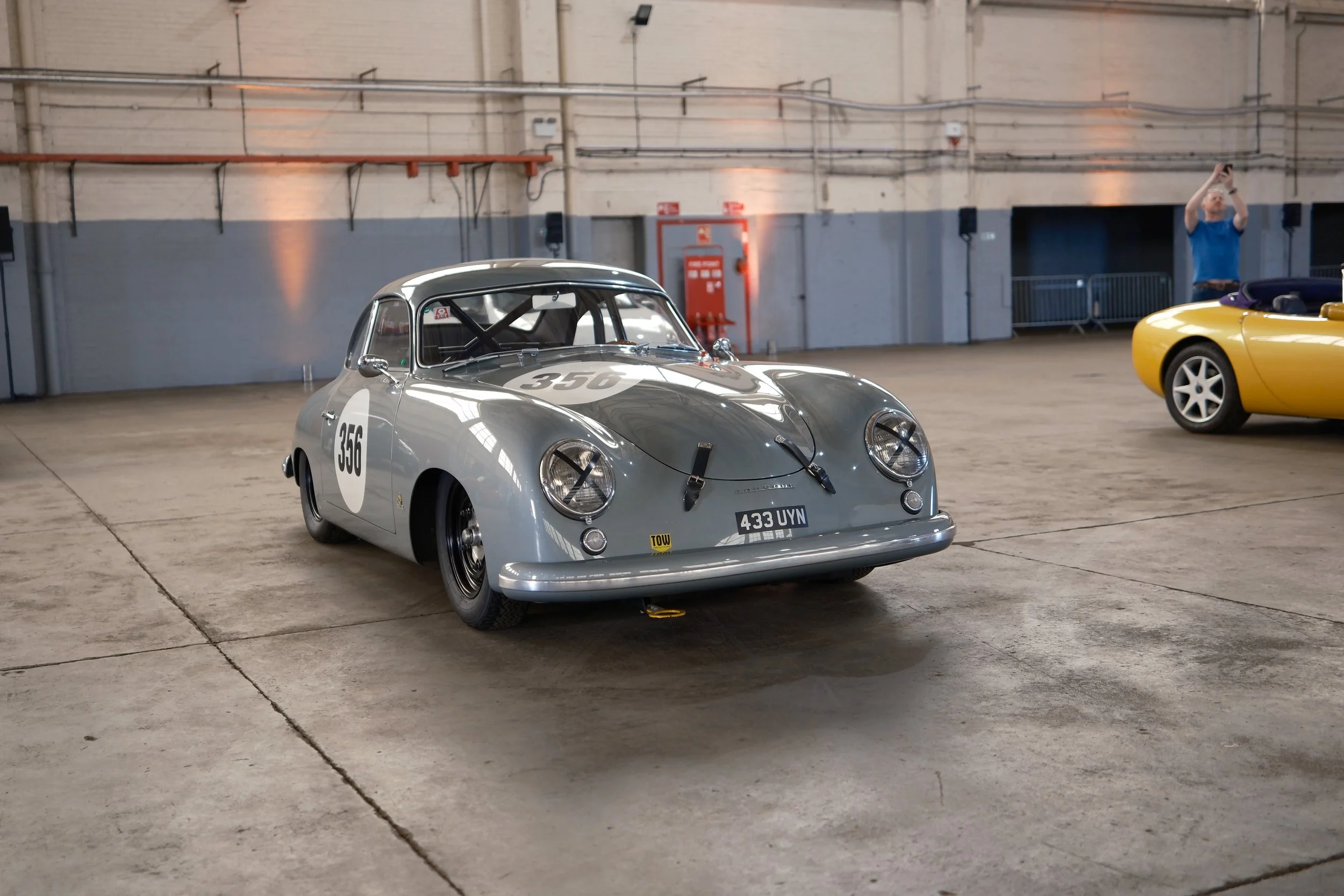 A vintage race car with the number 356 and a rear license plate 433 UYN parked inside a large industrial warehouse, with a yellow convertible sports car partially visible in the background and a person taking a photo.