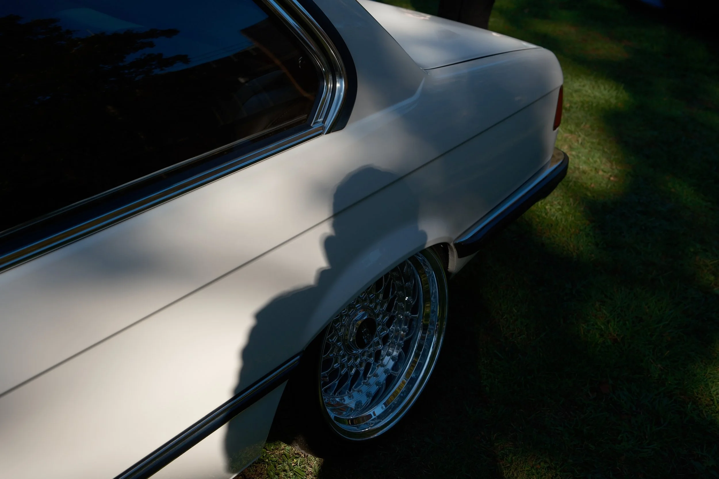 Close-up of the rear side of a classic silver car with spoked wheels, parked on grass, with shadows cast on the car body.