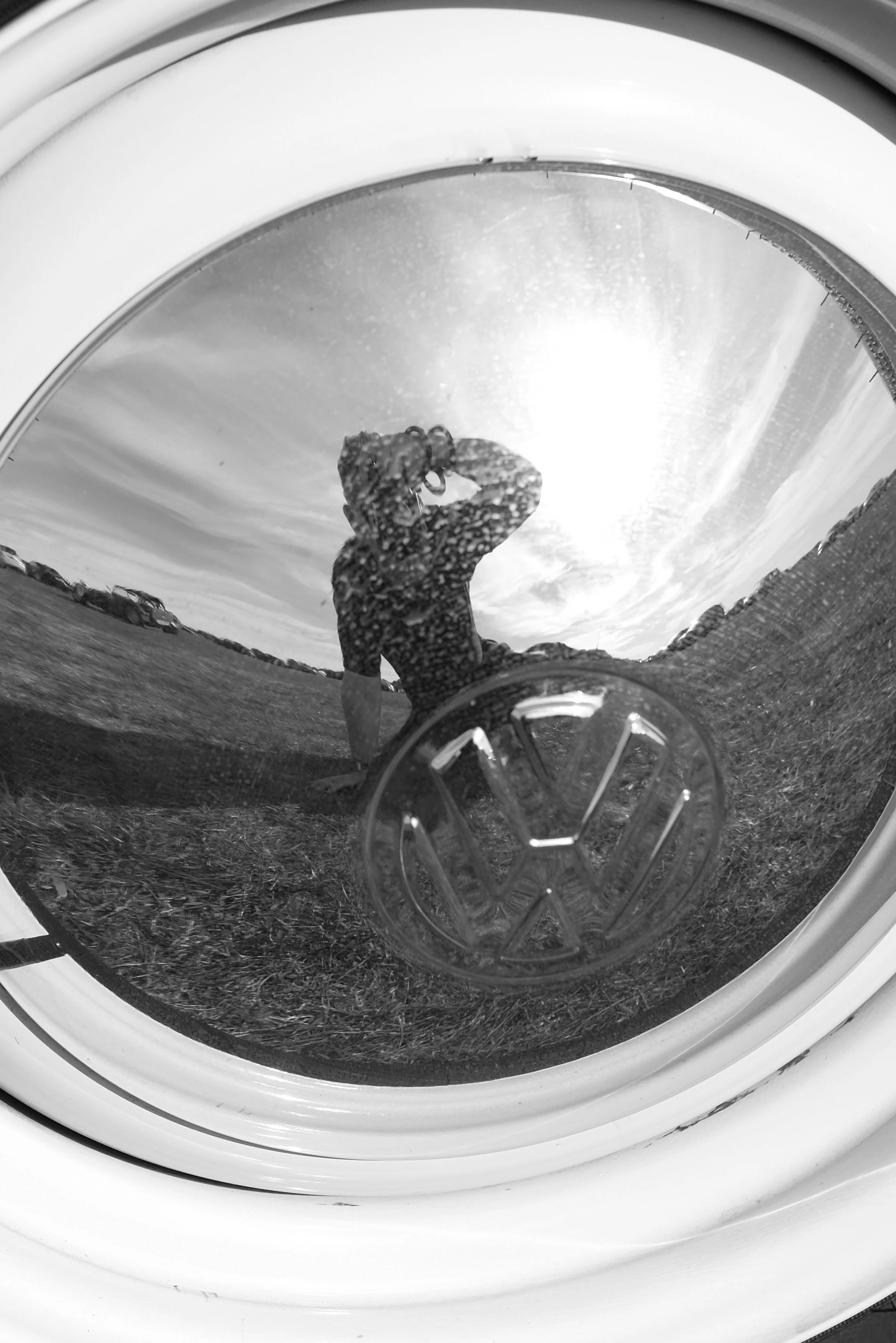 Reflection of a person taking a picture in a washing machine drum, with a cloudy sky in the background.
