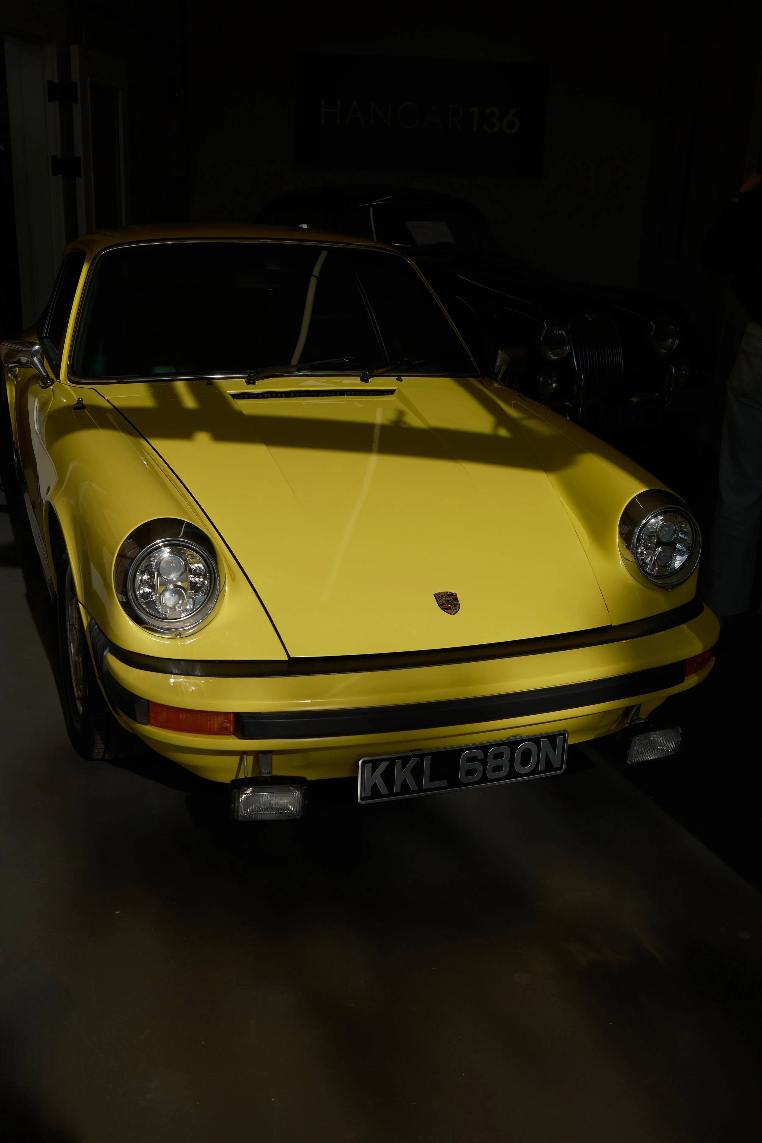 A yellow vintage Porsche 911 sports car with a black license plate reading 'KKL 680N', displayed indoors with a dim background and a sign that reads 'HANGAR 136'.
