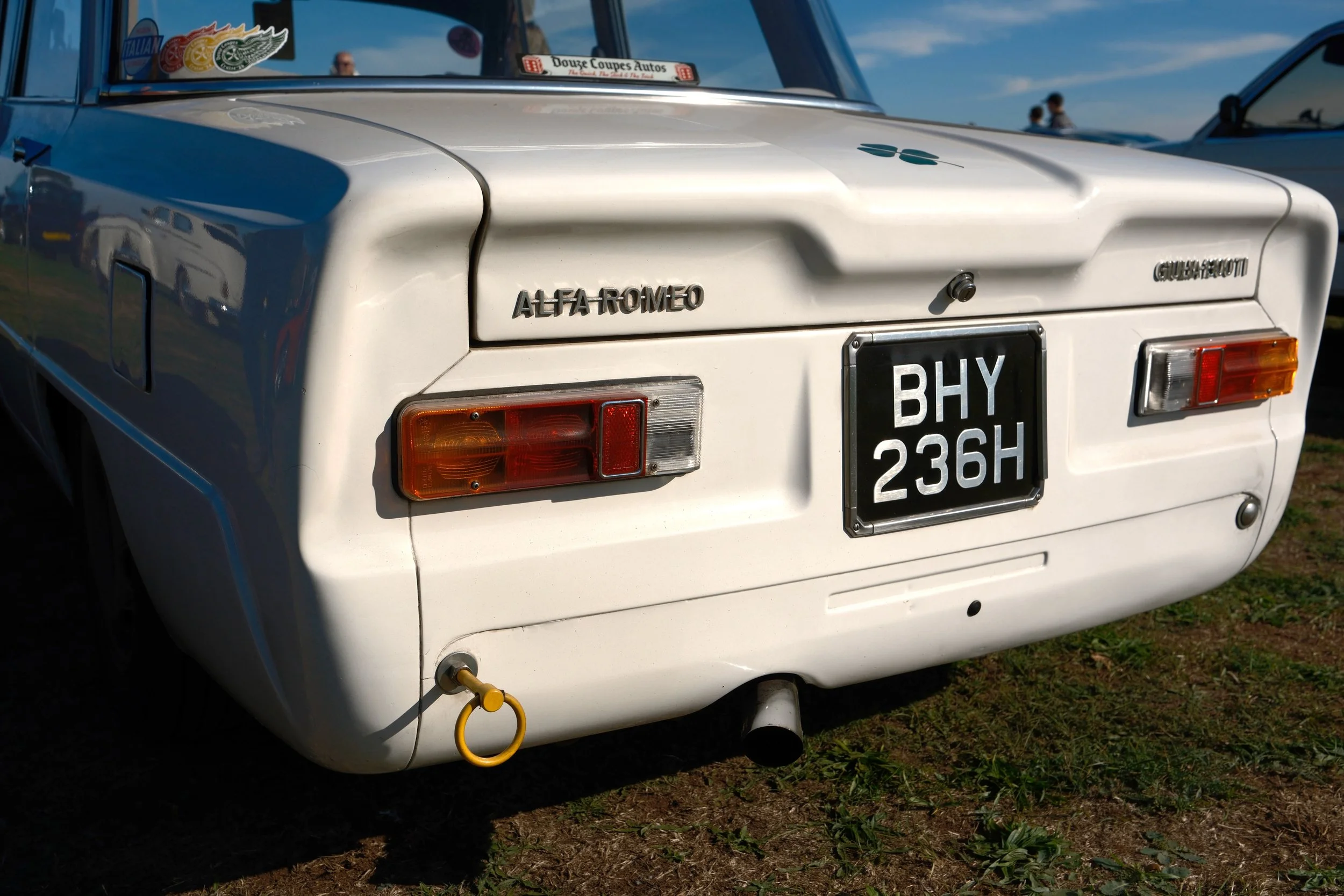 Rear view of a white classic Alfa Romeo car with a black license plate reading "BHY 236H," parked on grass. A yellow cable is connected to the back of the car. The car has tail lights and a badge that reads "Alfa Romeo," and the front part shows part