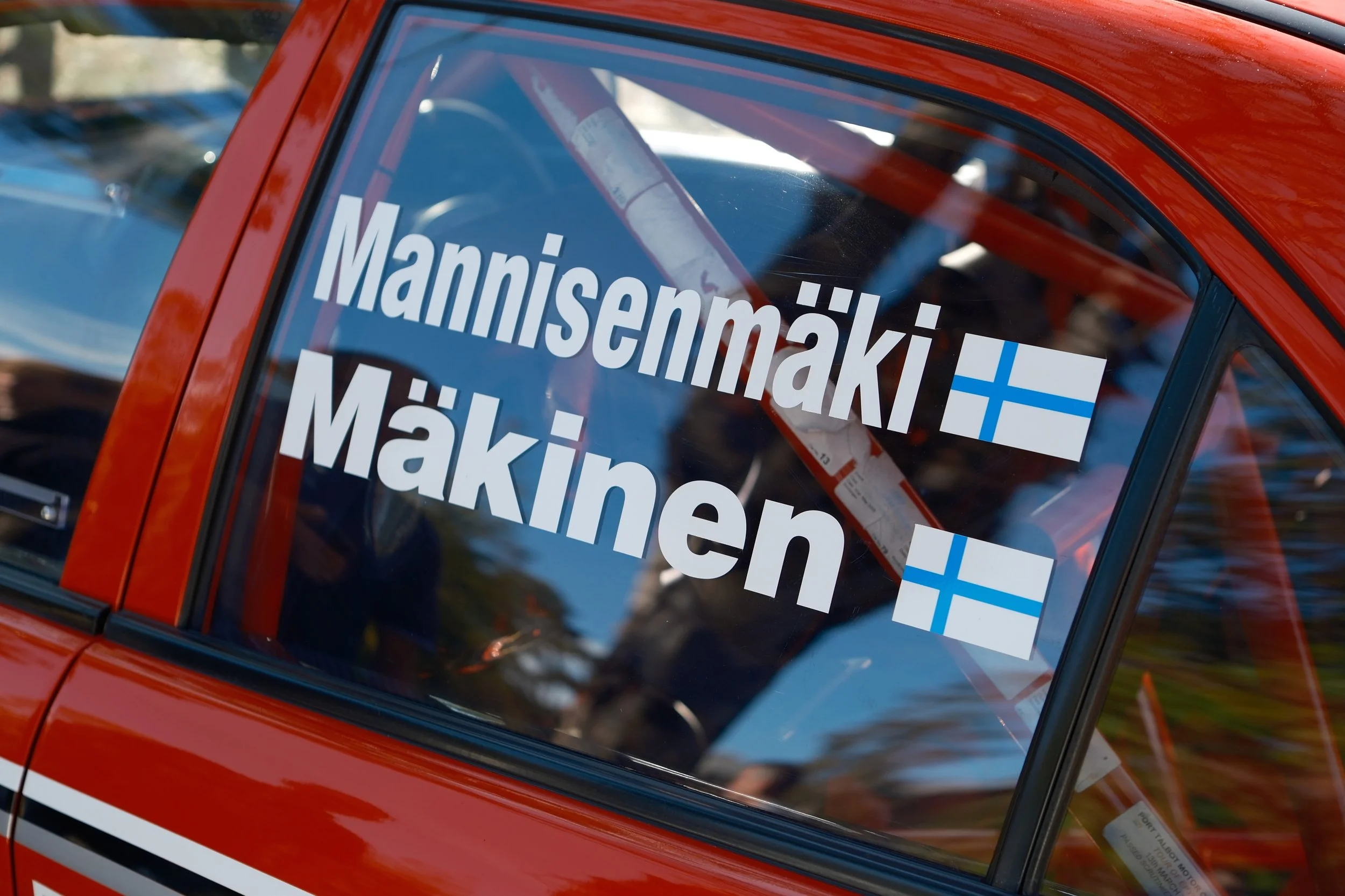 Close-up of a red vehicle with a window displaying Finnish flags and the name Männistömäki. The vehicle has a racing or rally car appearance.