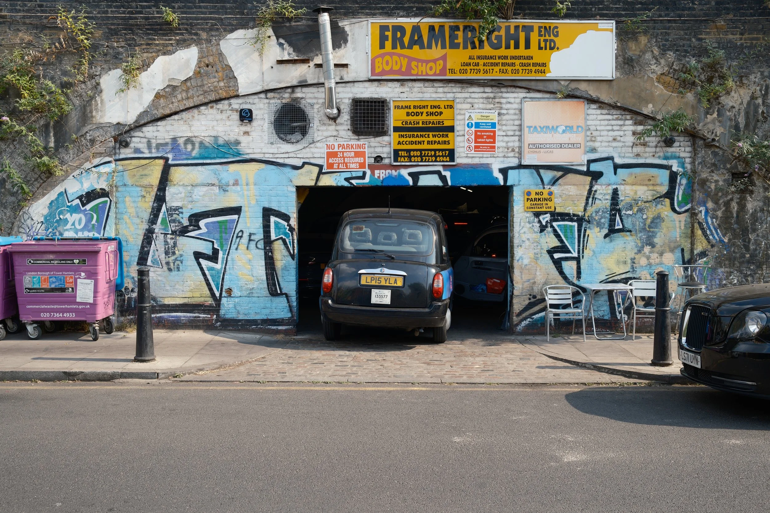 The image shows a graffiti-covered building with multiple signs above a garage entrance. There are two cars parked in front and various chairs and a pink recycling bin on the sidewalk. The signs advertise body shop, crash repairs, insurance work, and