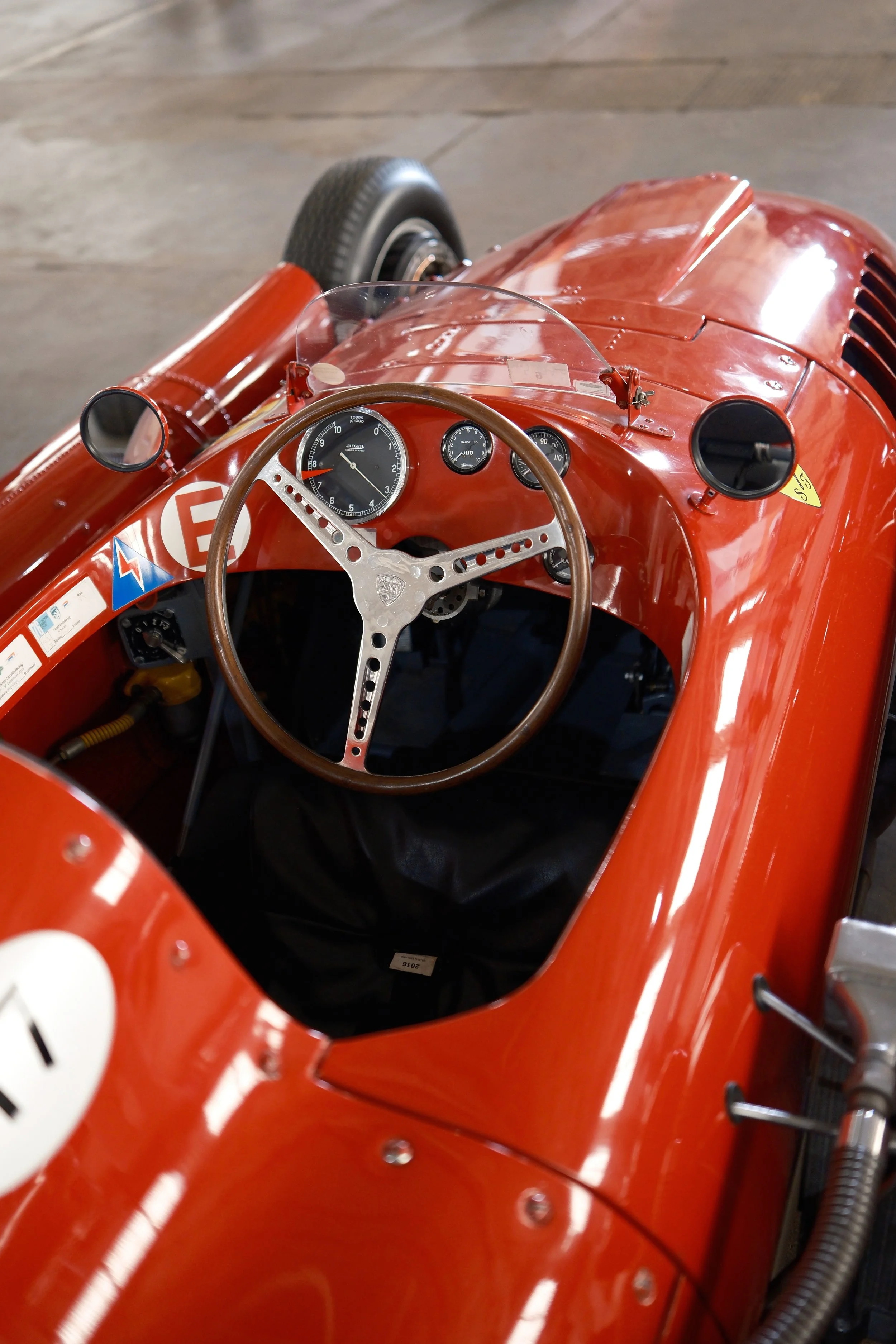 Red vintage race car dashboard with gauges, steering wheel, mirrors, and hood, viewed from the driver's seat.