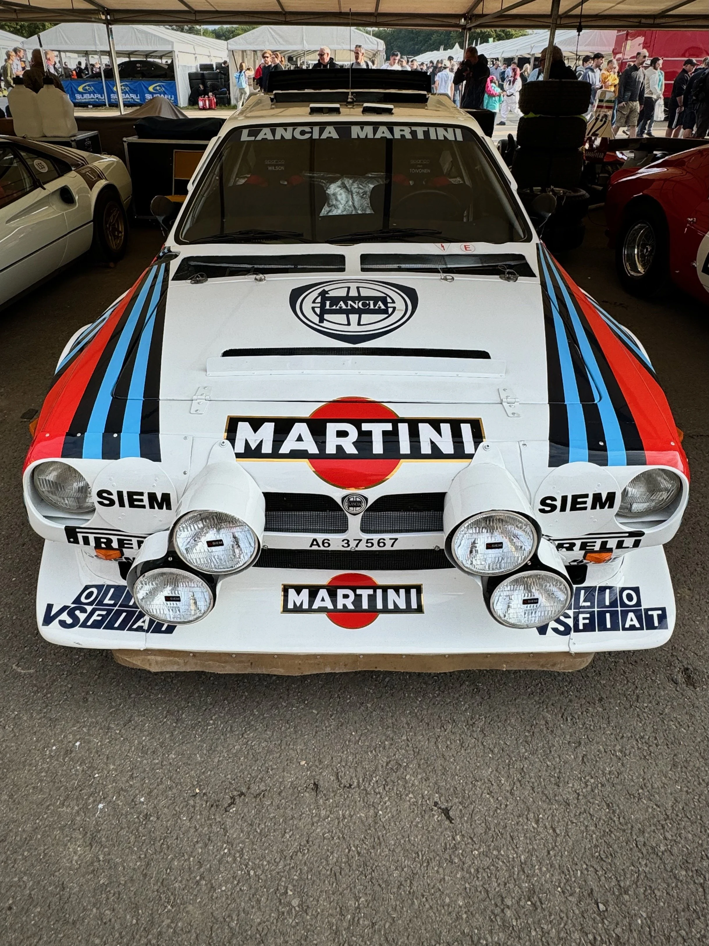 Front view of a vintage Lancia rally car with Martini racing livery, multiple auxiliary lights, and sponsorship decals, parked at a car show under a tent.