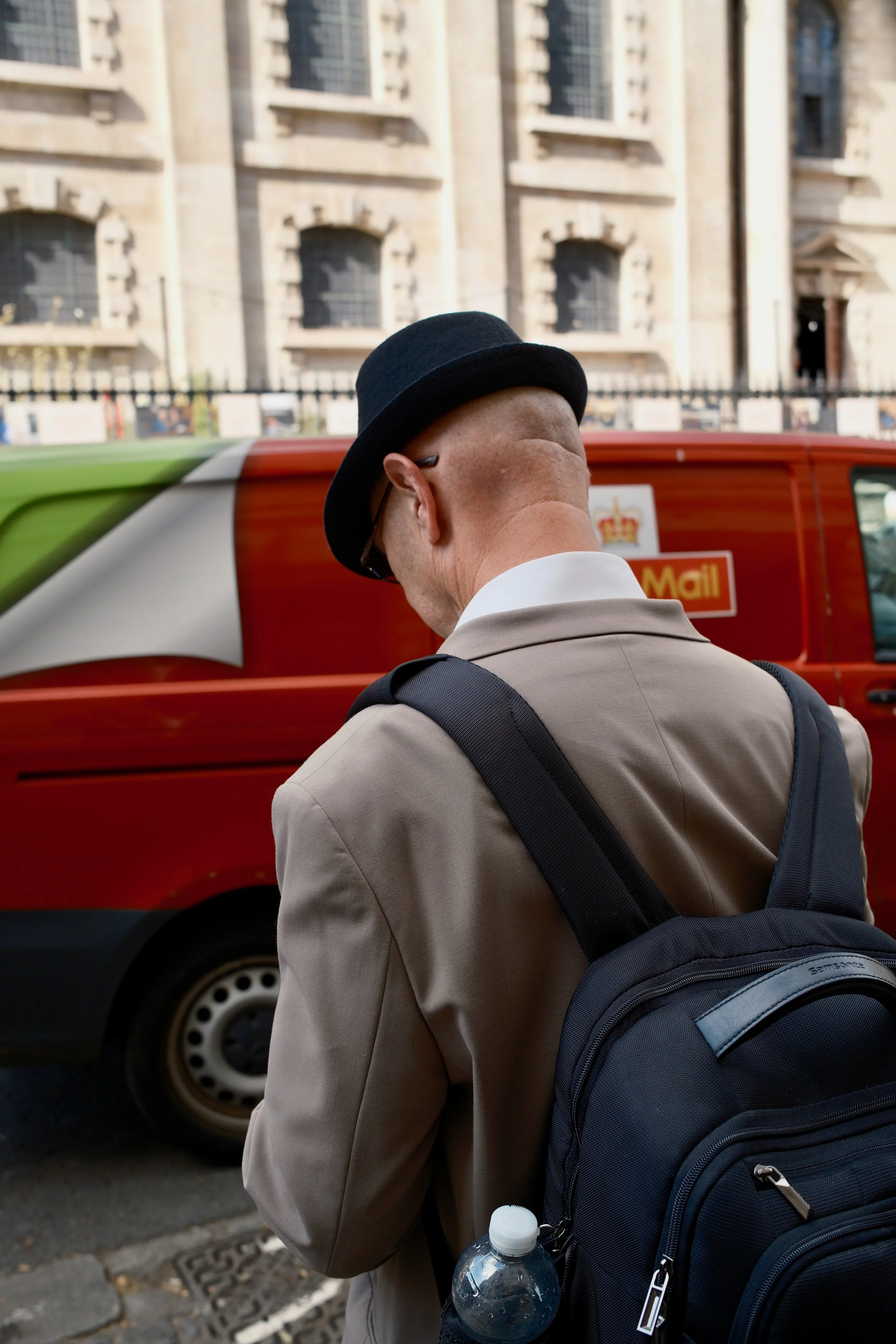 A man wearing a black hat and beige suit with a black backpack holding a water bottle is standing on a city street near a red mail truck, with old stone buildings in the background.