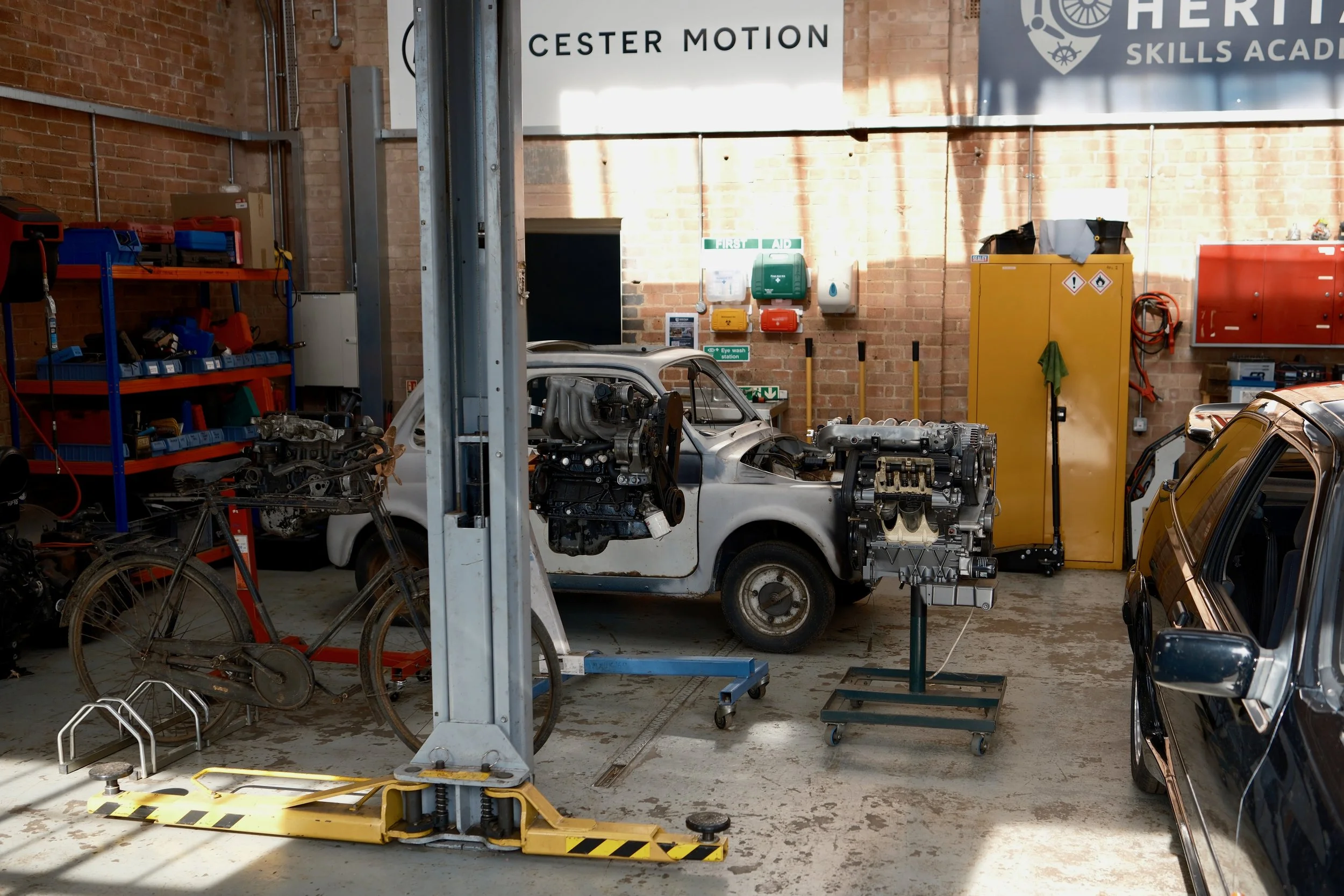 Inside a garage with brick walls, various tools, car parts, and machinery including a partially disassembled white car and a bicycle.
