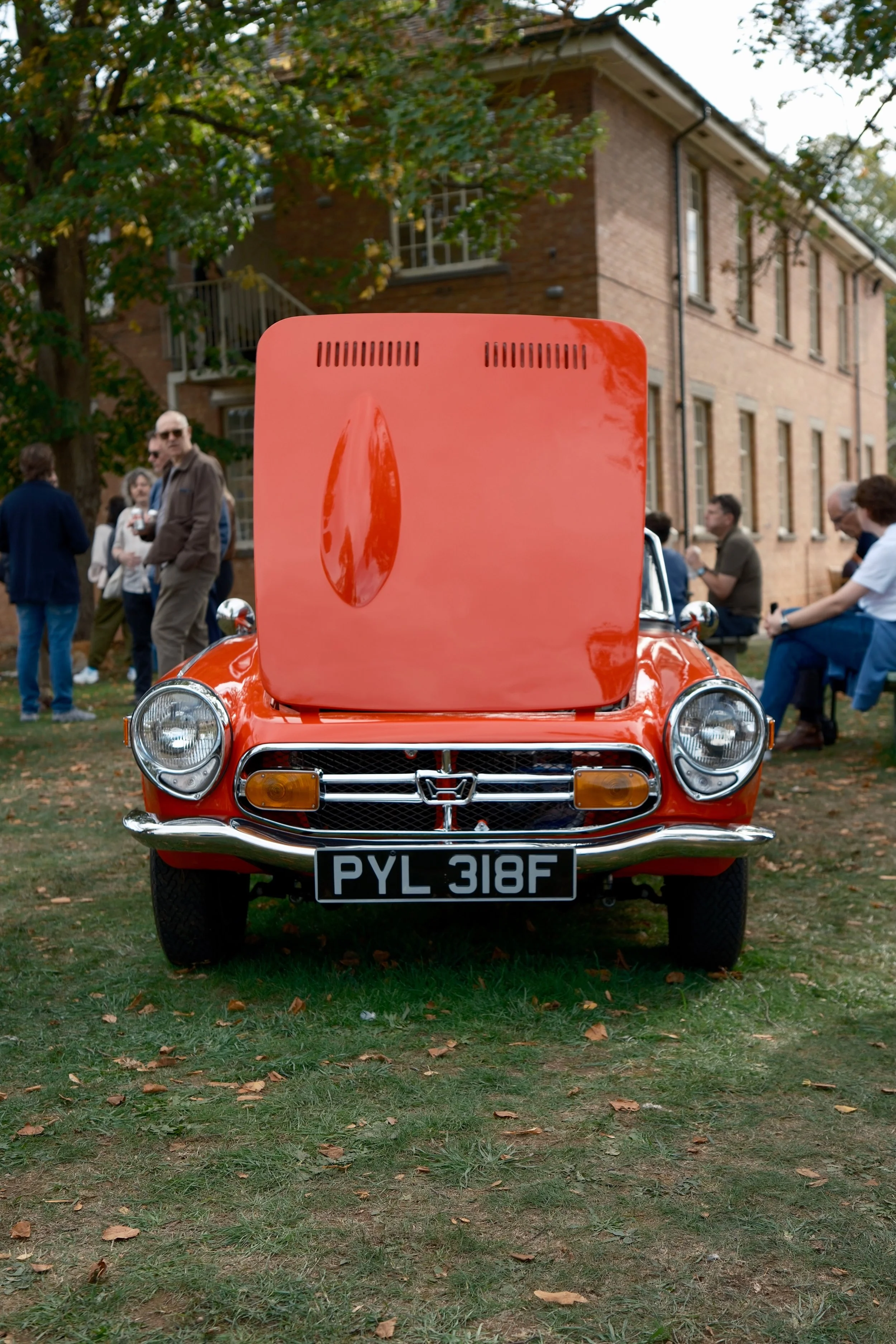 A vintage red-orange car with a raised hood, parked on grass with autumn leaves. In the background, there are people mingling and talking, and a brick building with trees surrounding it.