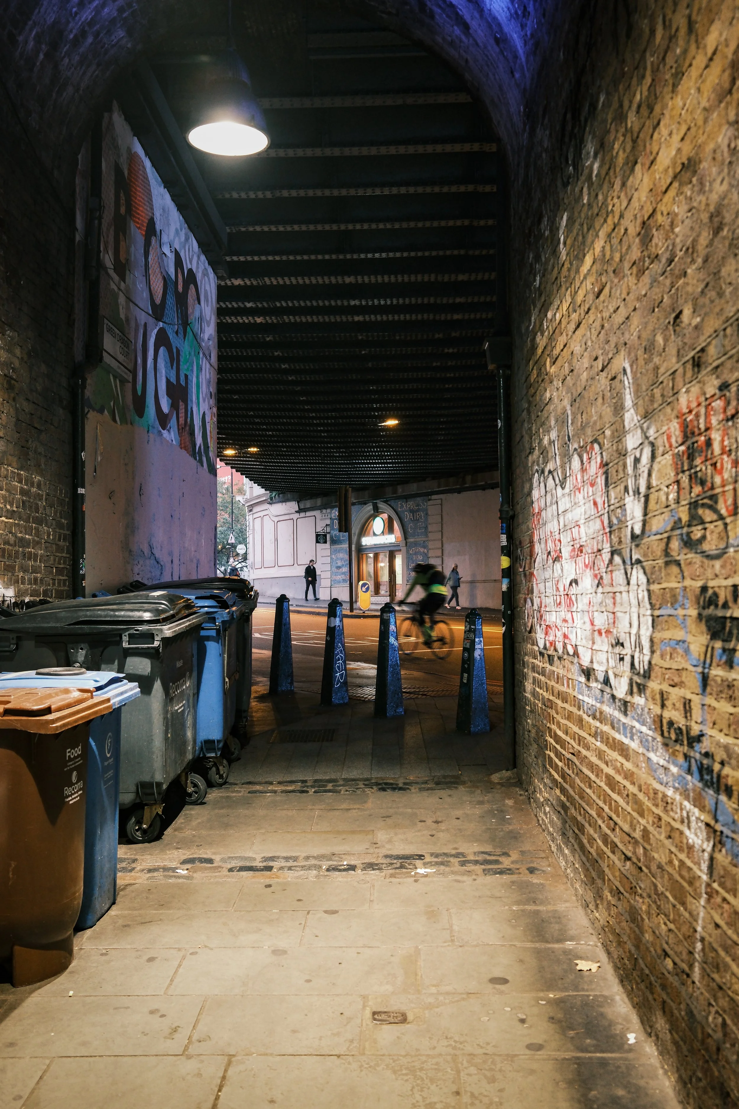 Underpass with brick walls on either side, a sidewalk in the foreground, large trash bins on the left, graffiti on the right wall, and a cyclist riding through the underpass towards a street with a building and pedestrians in the background.
