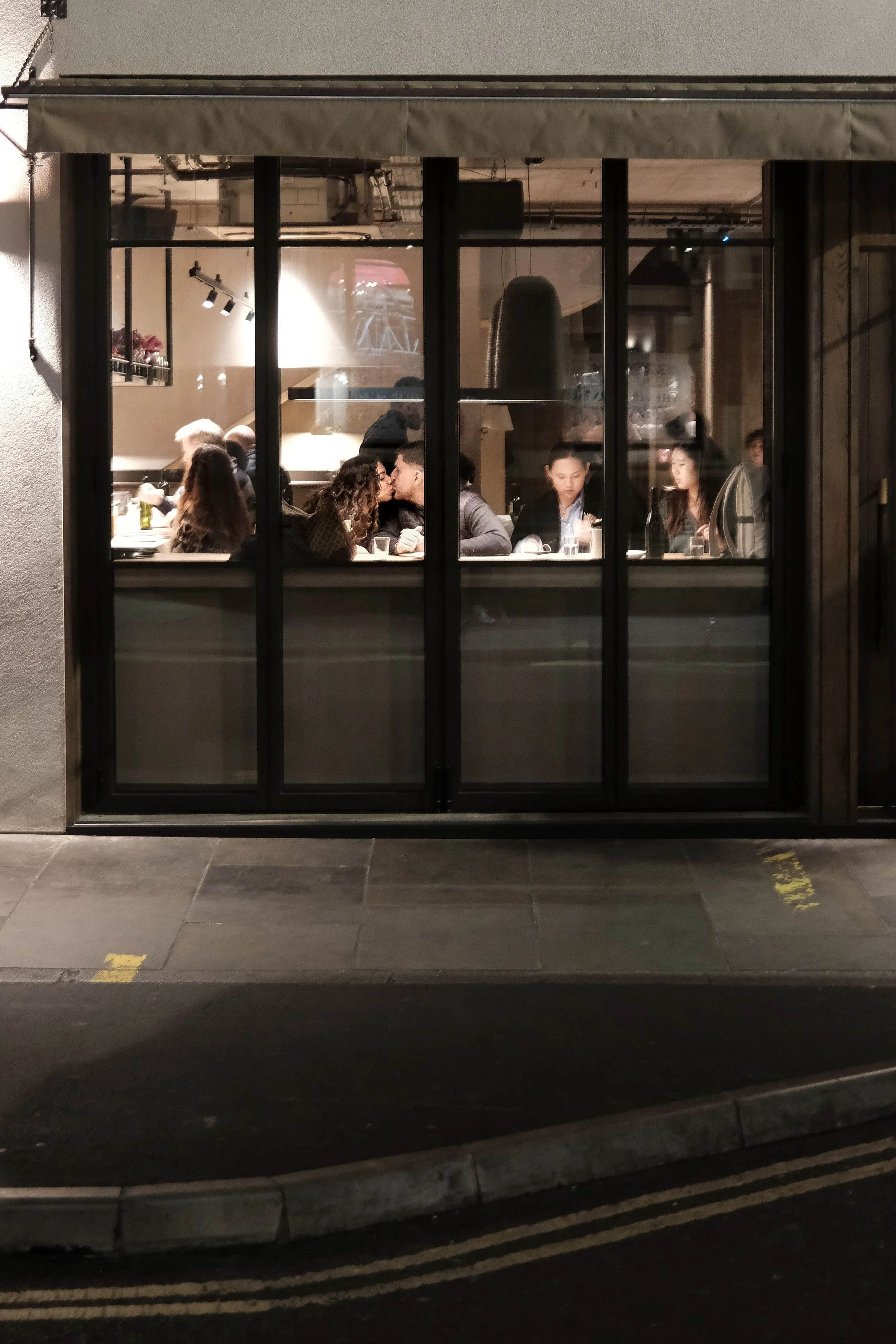 Nighttime view of a restaurant window with people sitting inside, including couples kissing and talking, with the street and sidewalk outside.