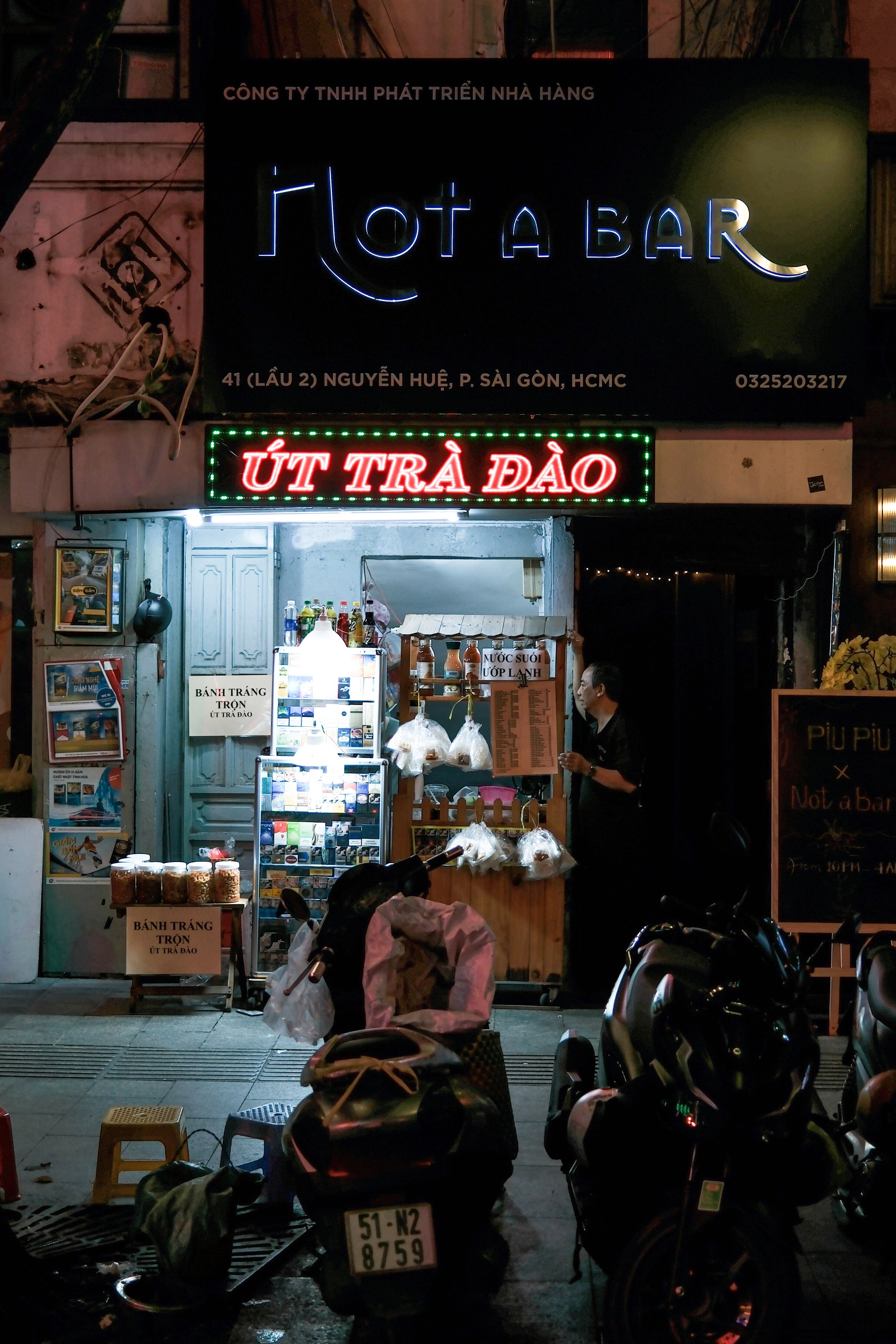 Nighttime street scene with a small food stall illuminated by neon and LED signs, selling bánh tráng trộn and other snacks, with parked motorbikes in front and a man attending the stall.