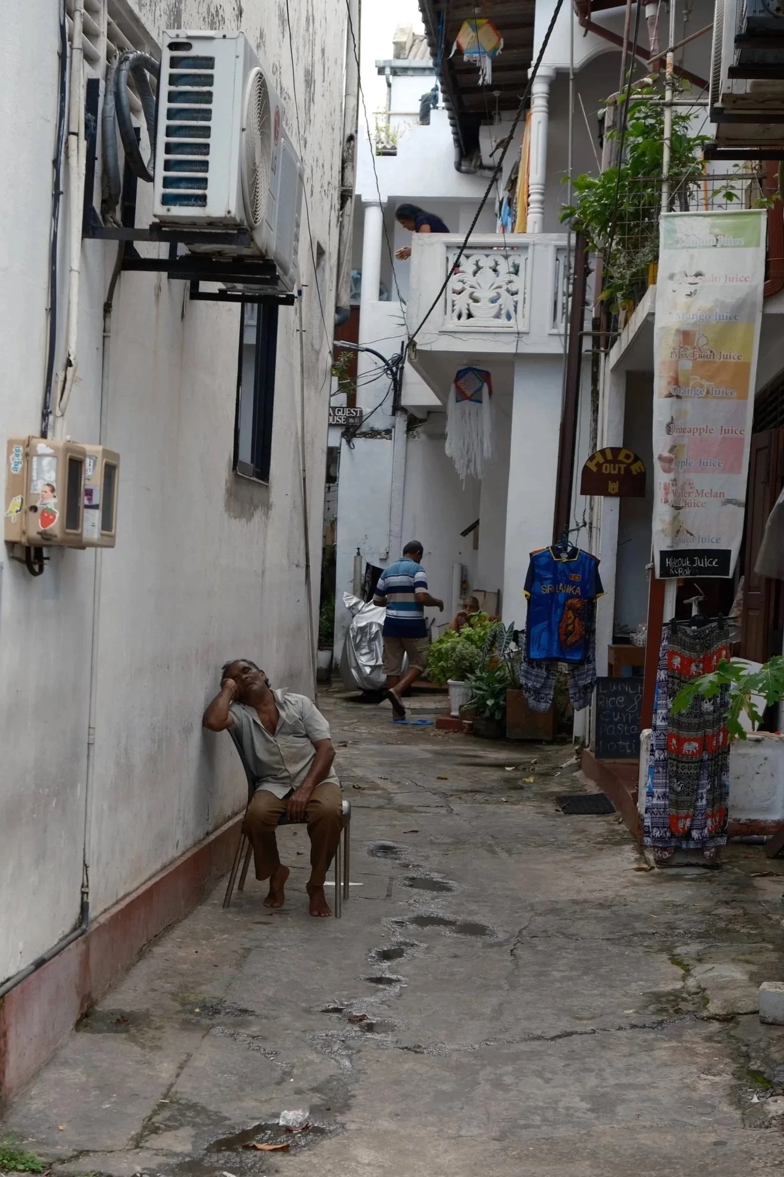 An alleyway with white buildings and various shops, including a juice stand, with a man sitting on a chair, a woman on a balcony, and another man walking.