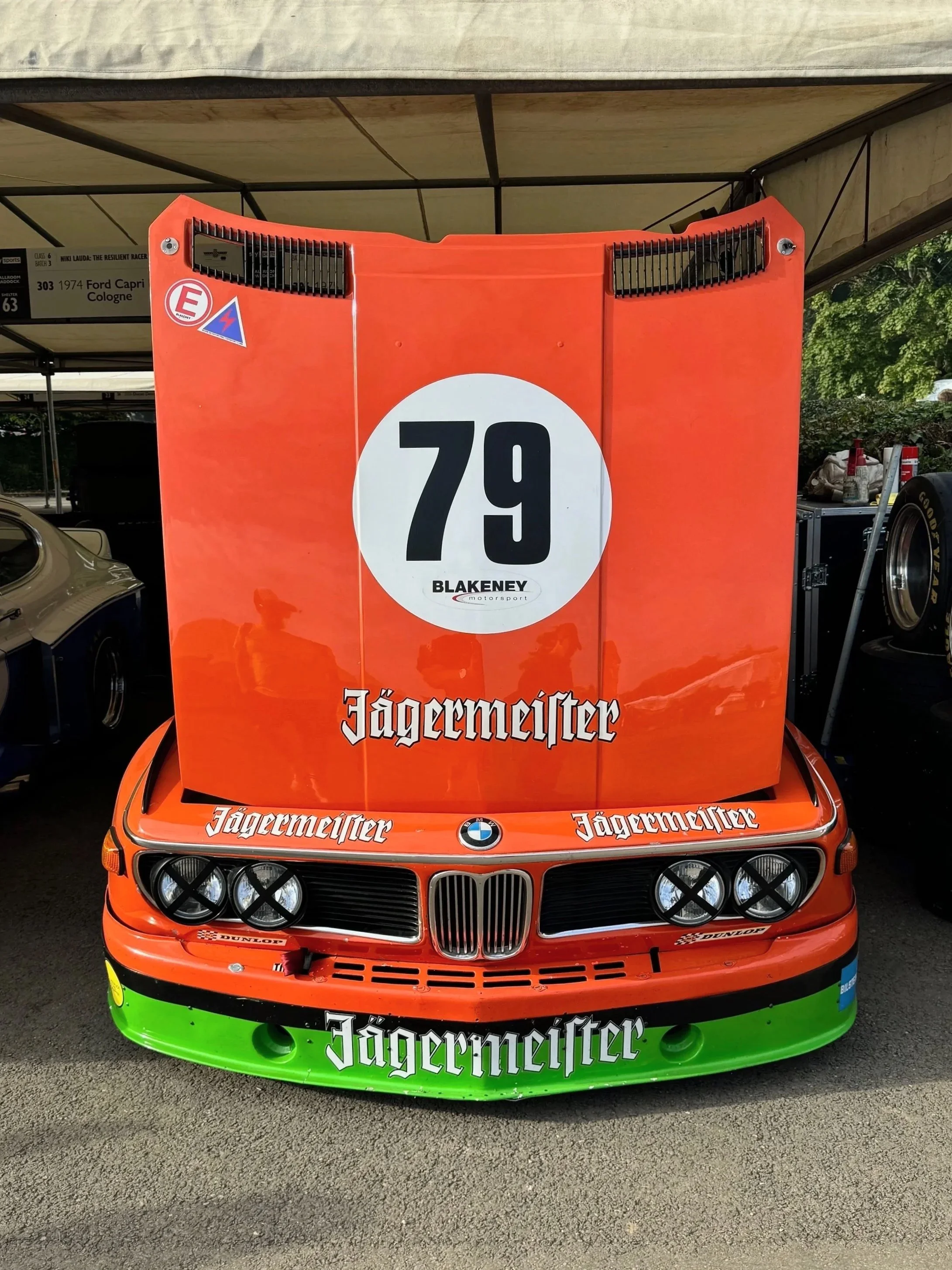 A vintage race car with orange and green Jägermeister branding, featuring a large white circle with the number 79 and the name Blakeney, parked in a garage area.