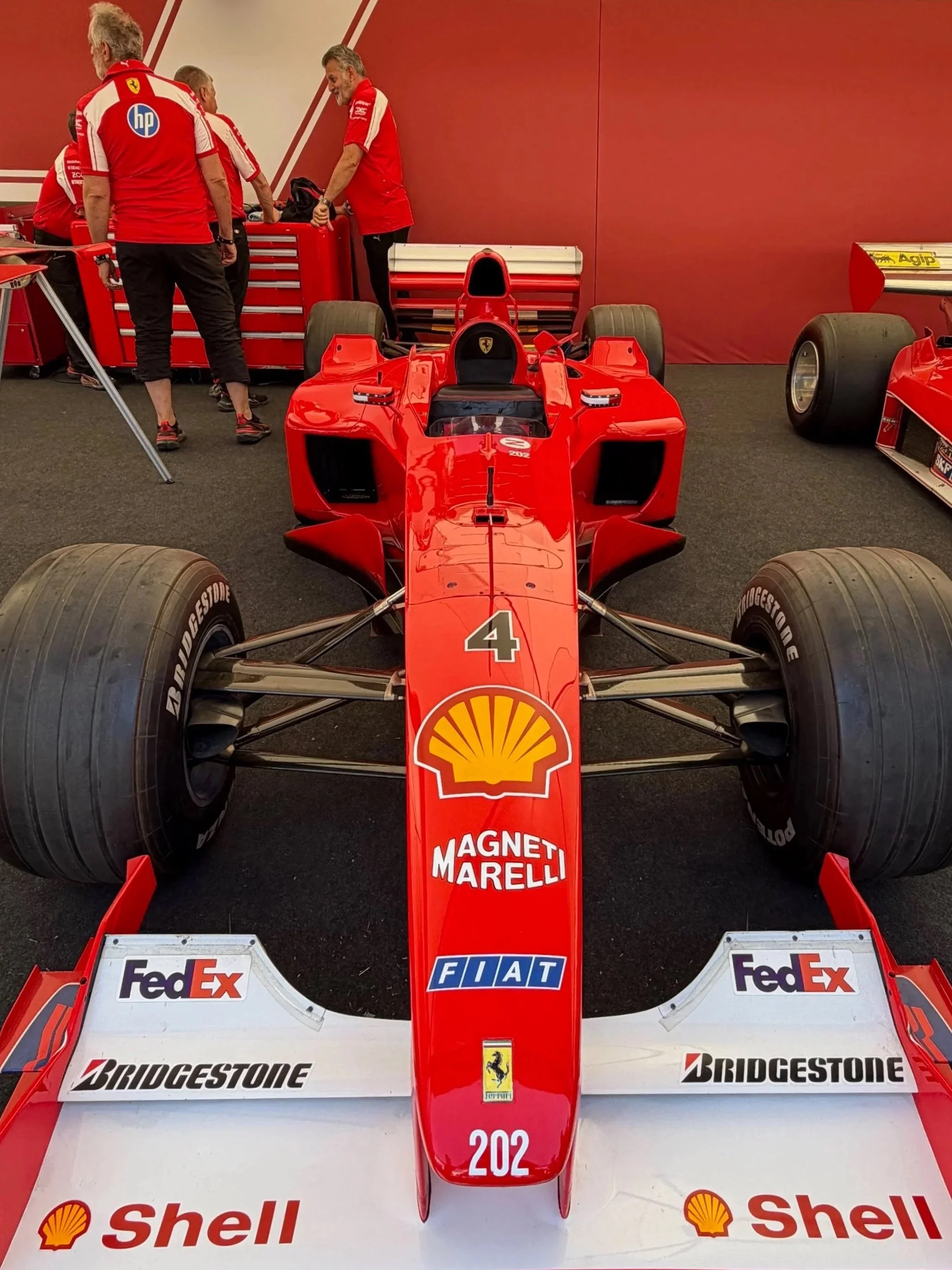 A vintage Formula 1 race car on display in a paddock area, with team members in red uniforms working and preparing the car. The car features sponsorship logos from Shell, Bridgestone, Shell, FedEx, FIAT, and Ferrari.