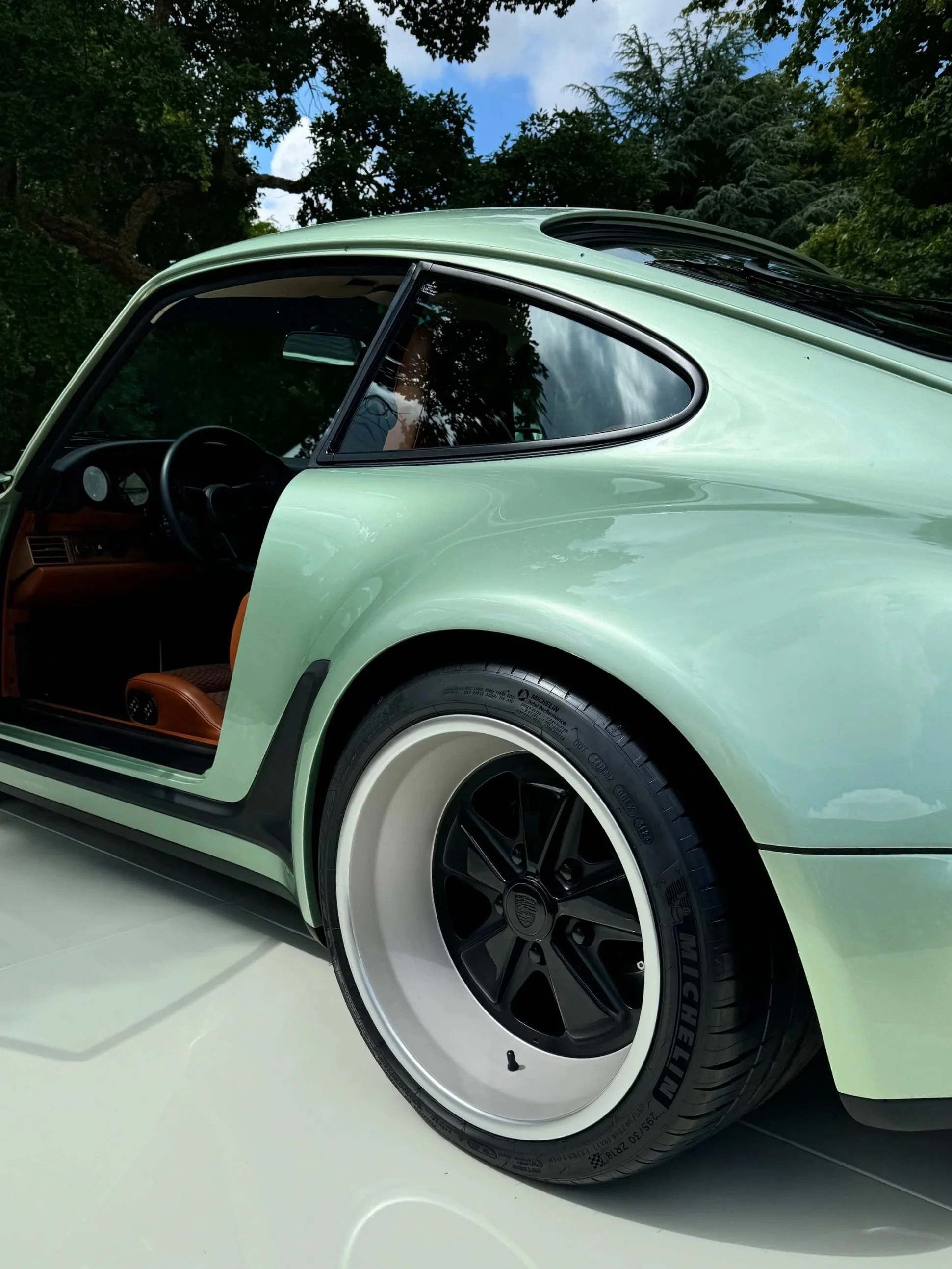 Close-up of a mint green vintage car with black wheels and whitewall tires, showing the side and partially open driver’s door, with reflections of trees and sky on the car's window.
