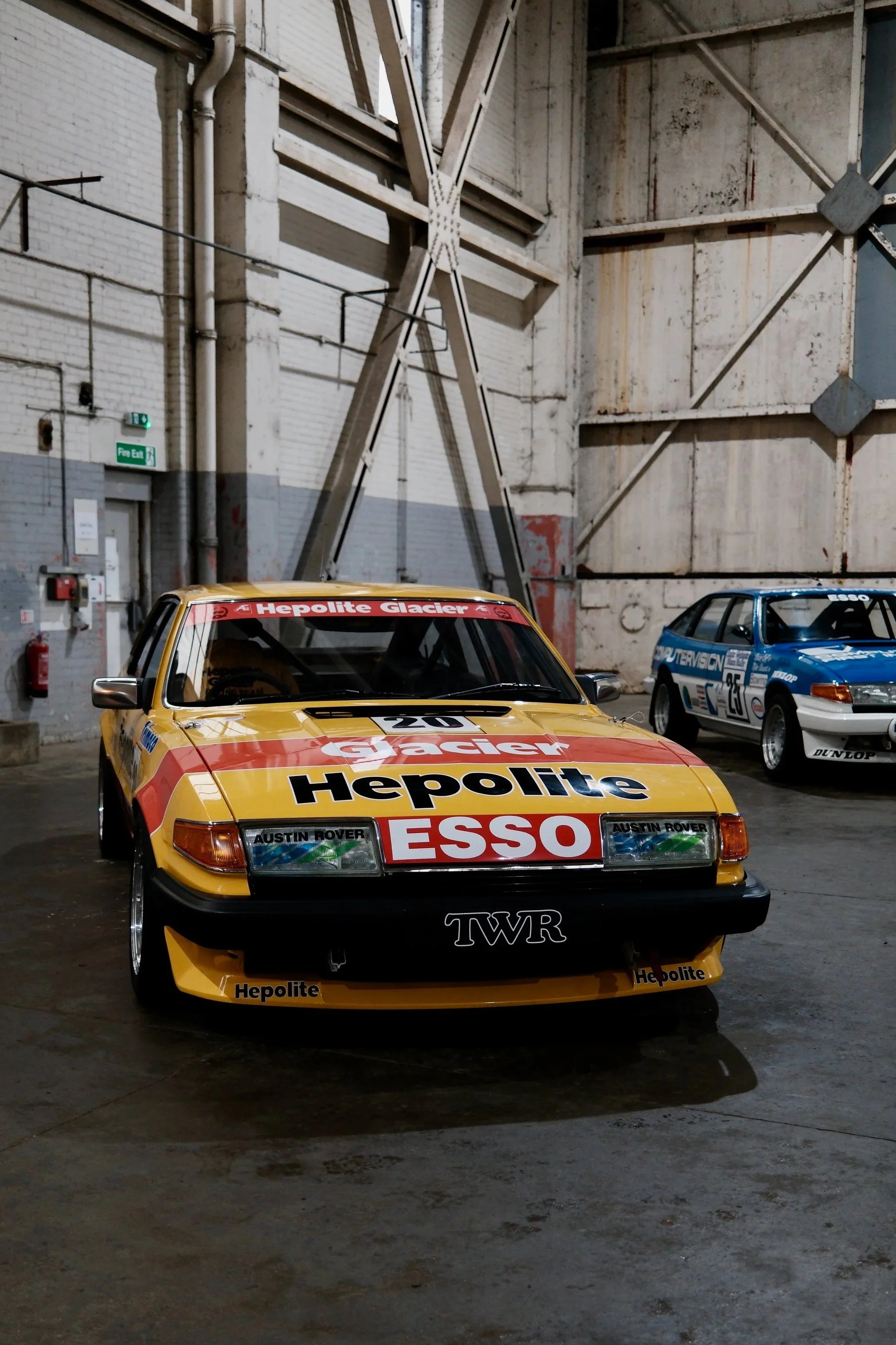 A yellow rally car with Kelpie and Esso sponsorships, parked inside a large industrial warehouse with white brick walls and large metal doors. Two other racing cars, one blue and one gray, are visible in the background.