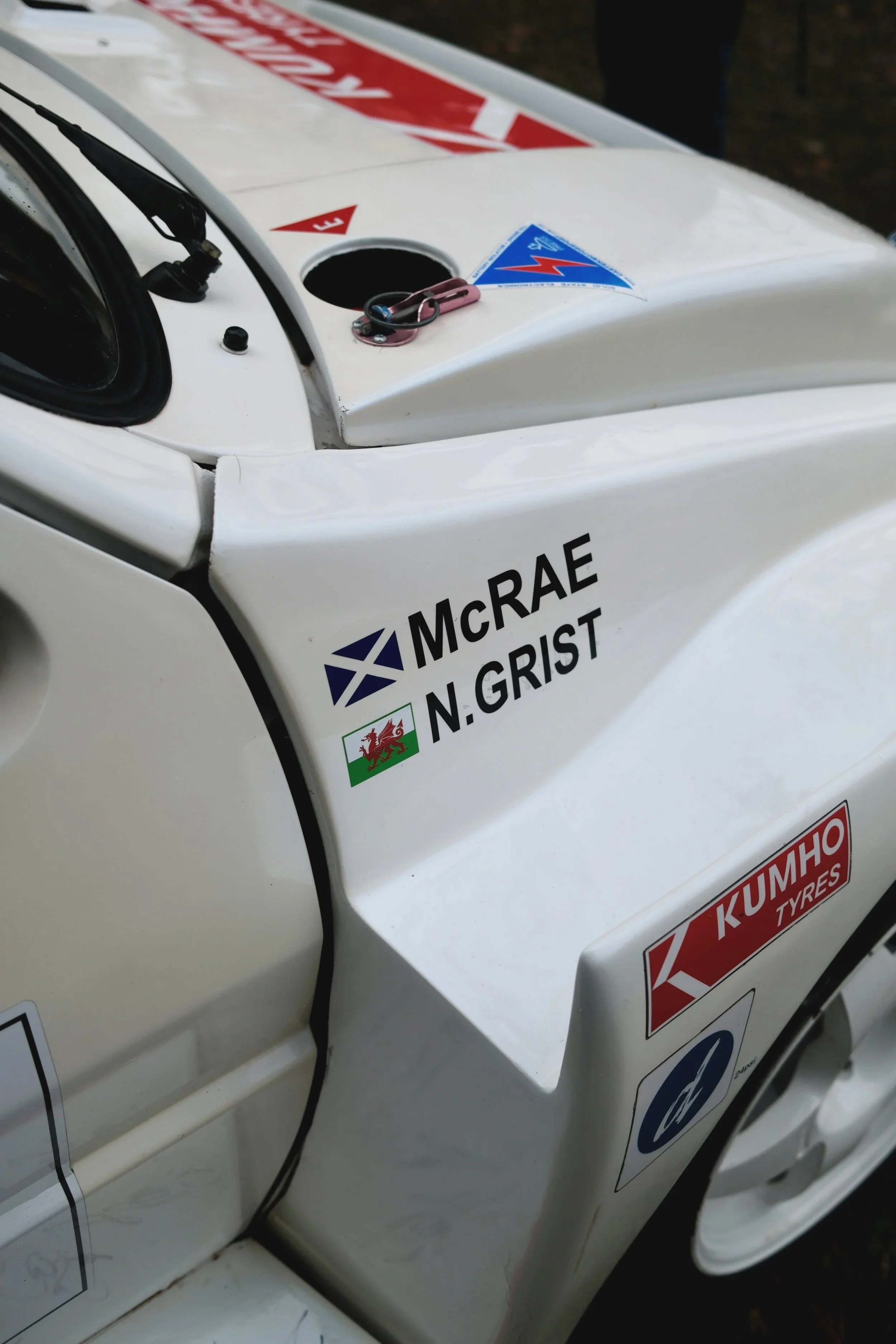 Close-up of a white racing car cockpit with sponsor decals, including McRae, N. Grist, KUMHO TIRES, and a Welsh flag, with a pink carabiner clip attached near the open fuel cap.