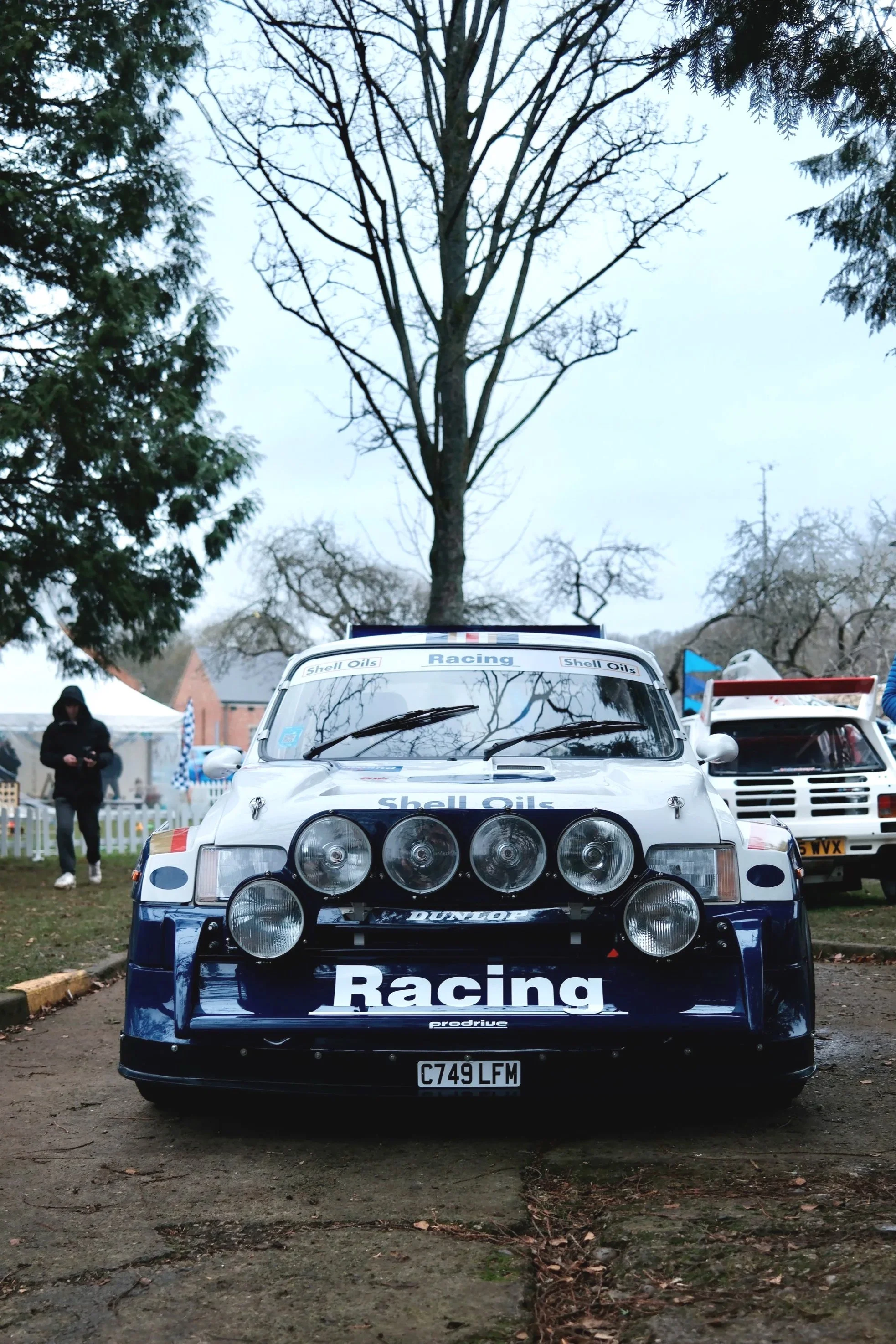 Front view of a classic rally car with sponsor decals, parked outdoors with trees and tents in the background.