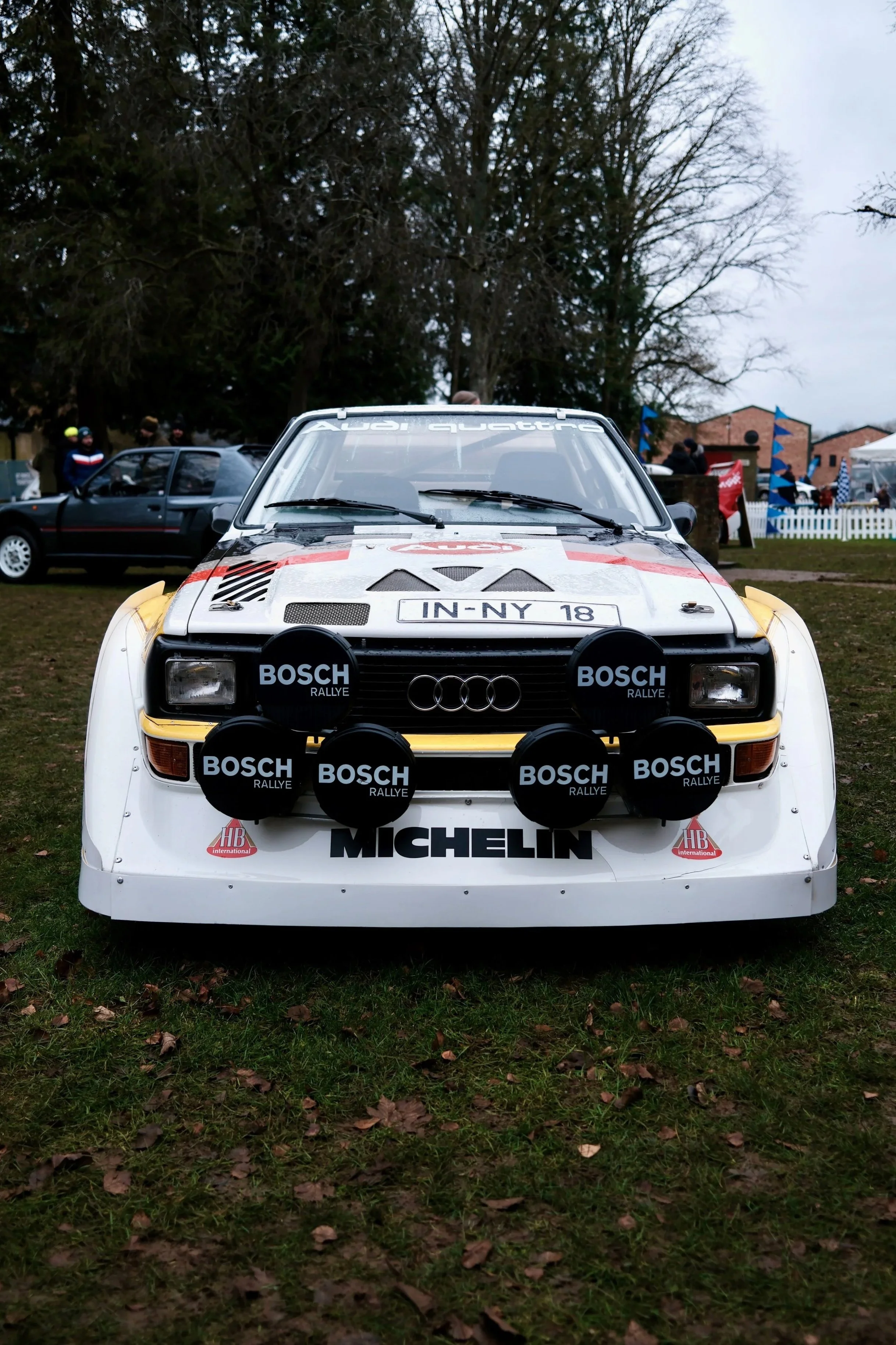 Classic white rally car with black and red sponsor decals, parked on a grassy field at an outdoor event with trees and buildings in the background.