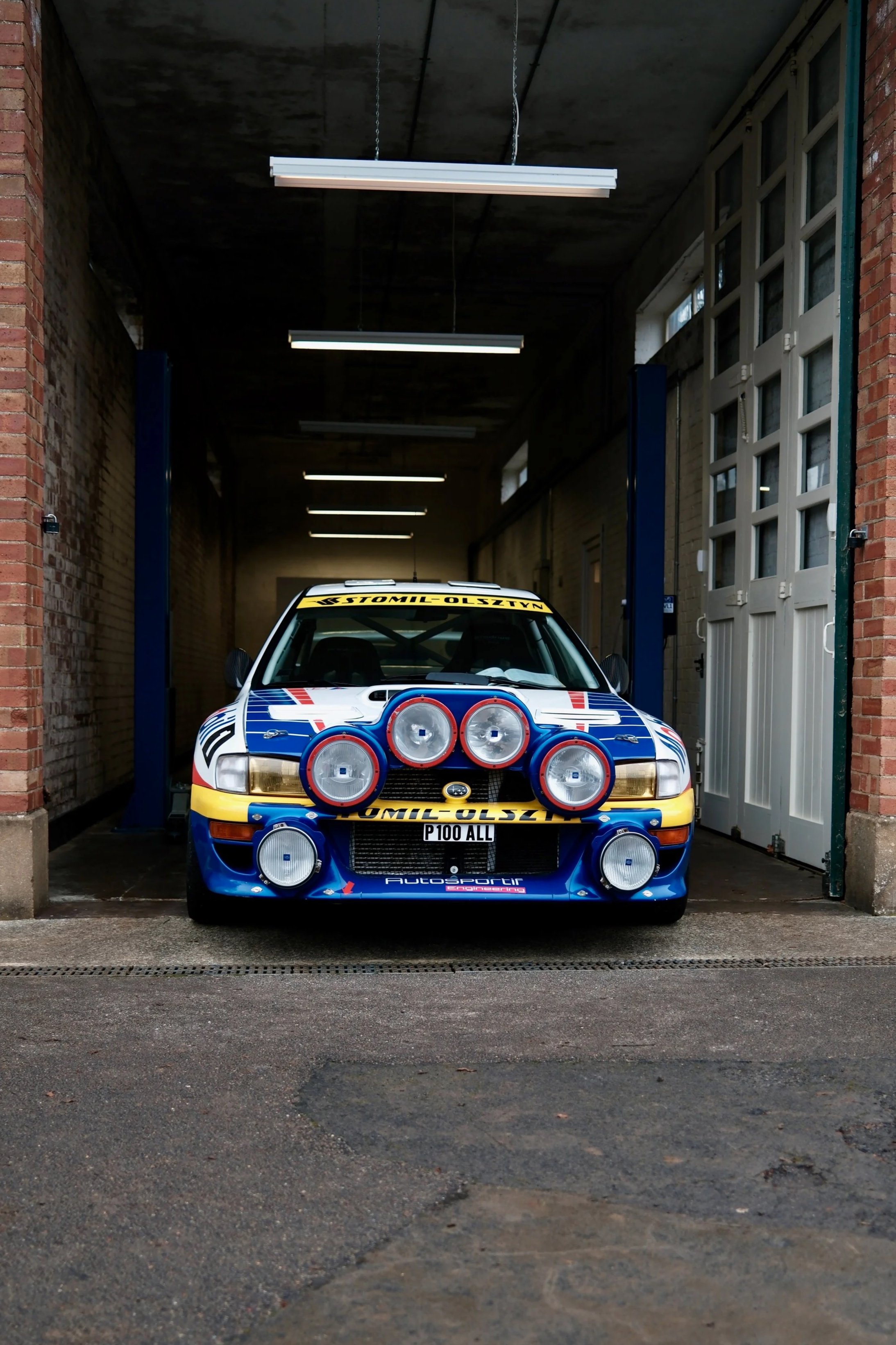 A rally car with multiple large spotlights mounted on the front, parked inside a garage with brick and metal walls and fluorescent lights overhead.