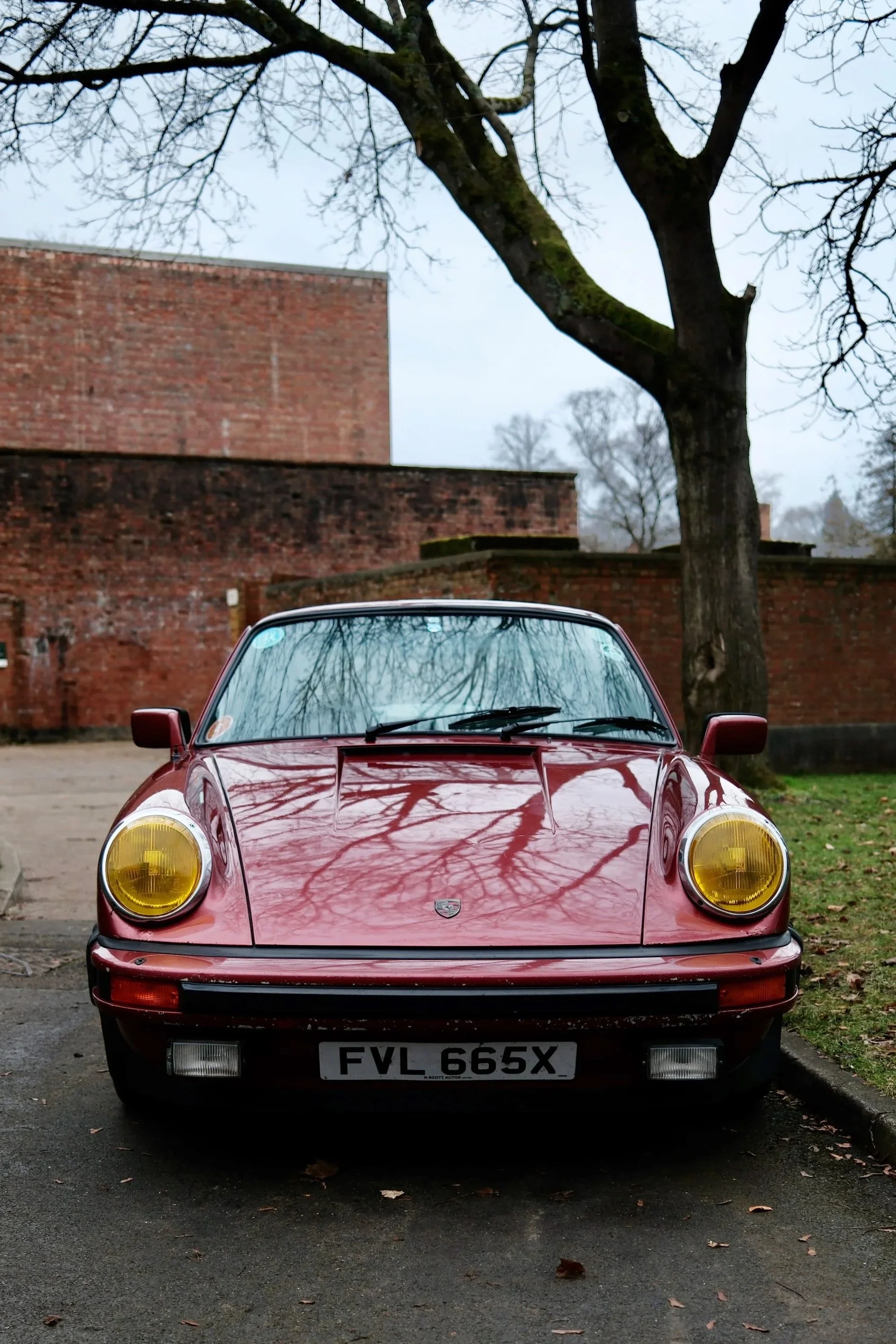 Front view of a red classic Porsche sports car with yellow fog lights, parked on a driveway with a brick wall, leafless trees, and an overcast sky in the background.