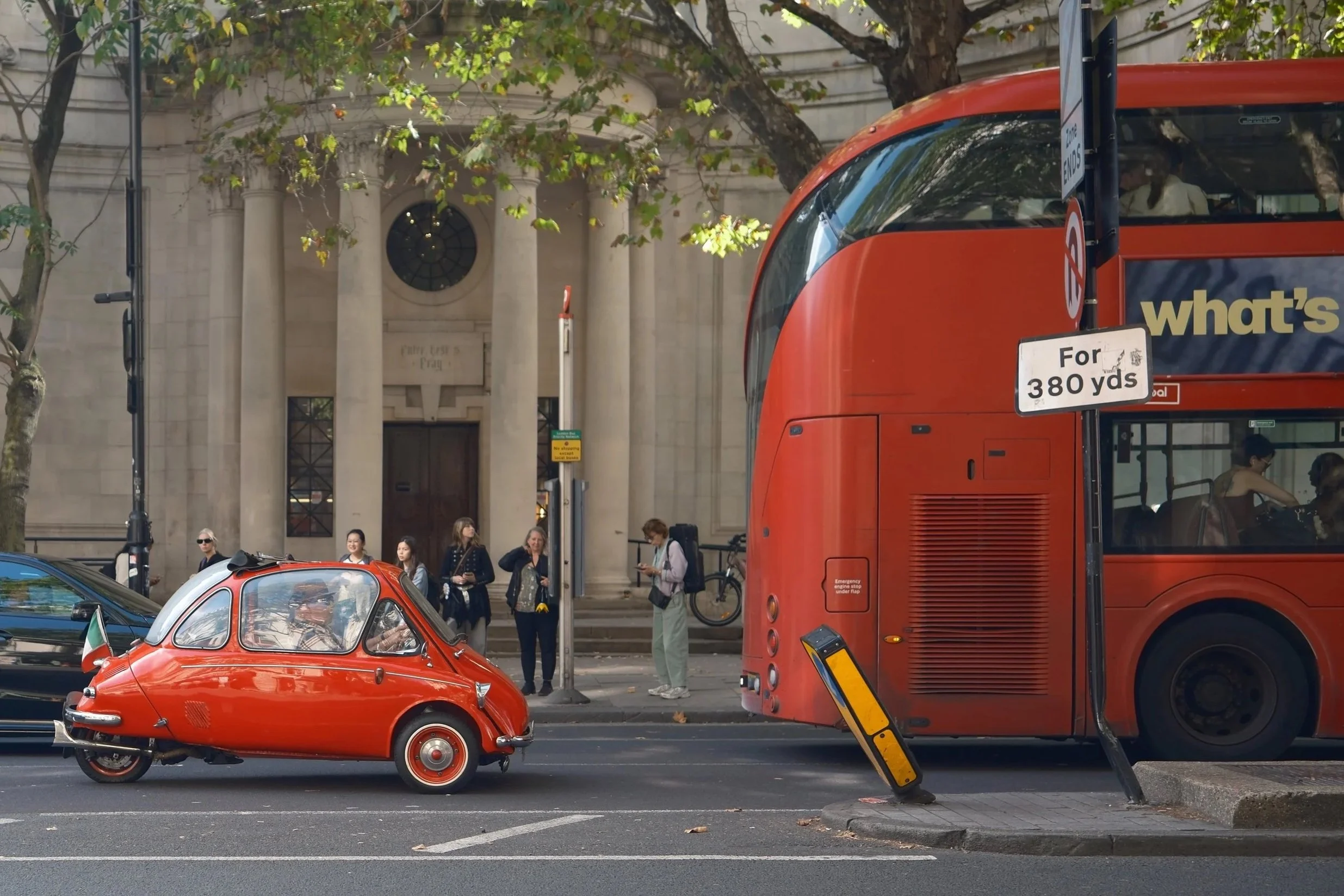 London street scene with a red double-decker bus, a classic red car, pedestrians waiting at the bus stop, and a historic building with columns in the background.