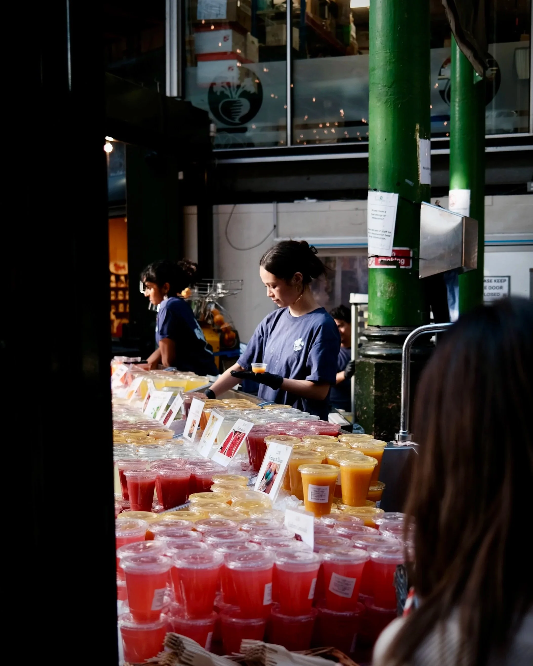 Various cups of colorful smoothies or juice on display at a market stall with indicators in front of each drink, and people in the background.