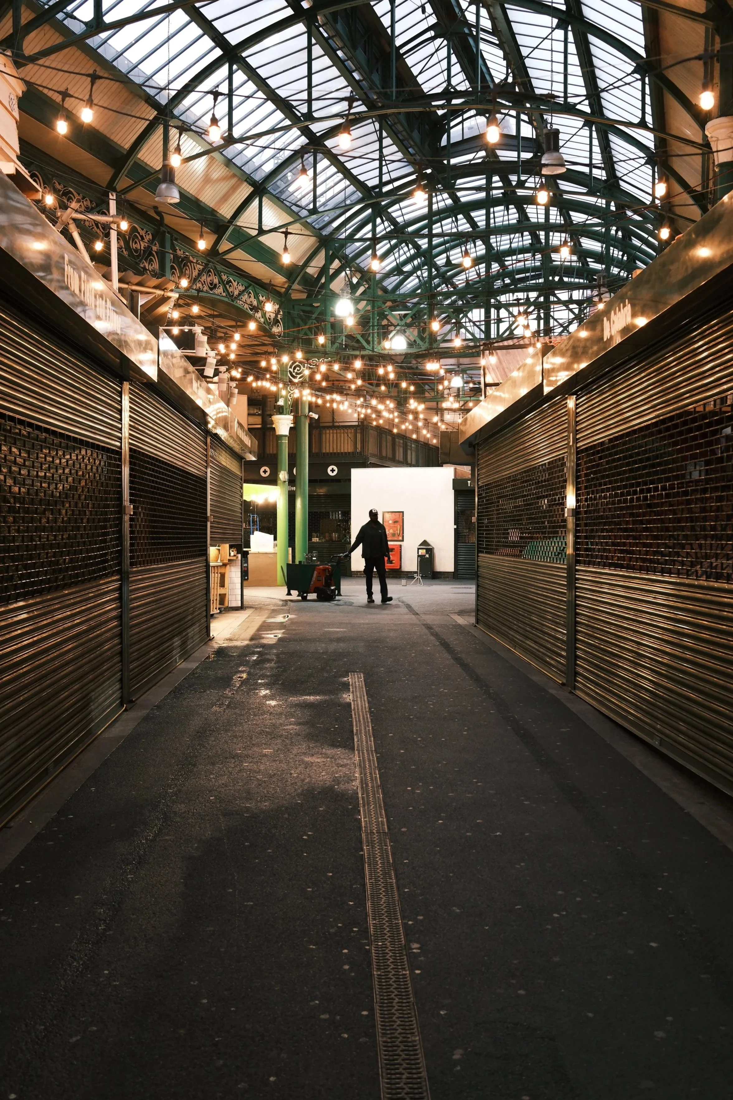An empty marketplace or shopping area with closed stalls, overhead string lights, and a glass ceiling. One person pushes a cart in the distance.