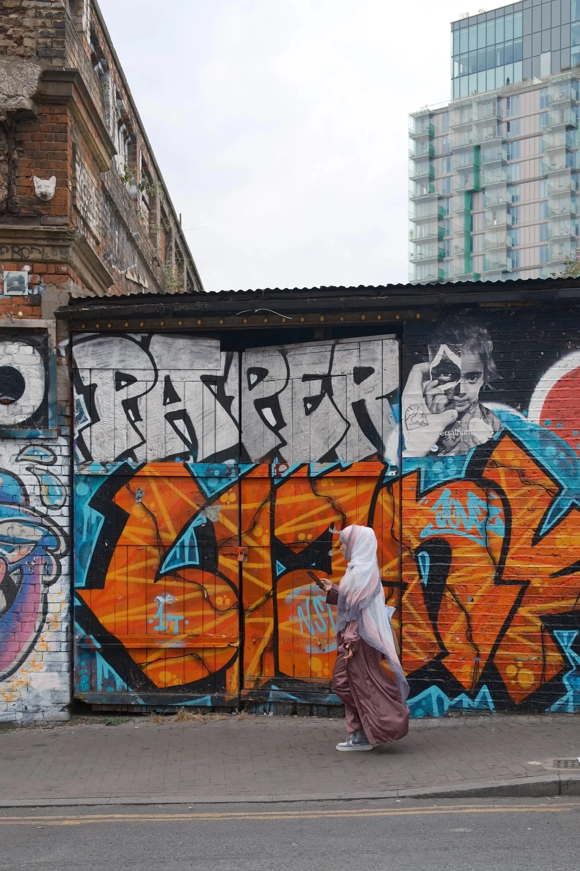 A person wearing a light purple hoodie and baggy pants walks past a colorful graffiti-covered wall. The scene takes place in front of an older brick building with a cat's face sculpture and a modern glass high-rise in the background.