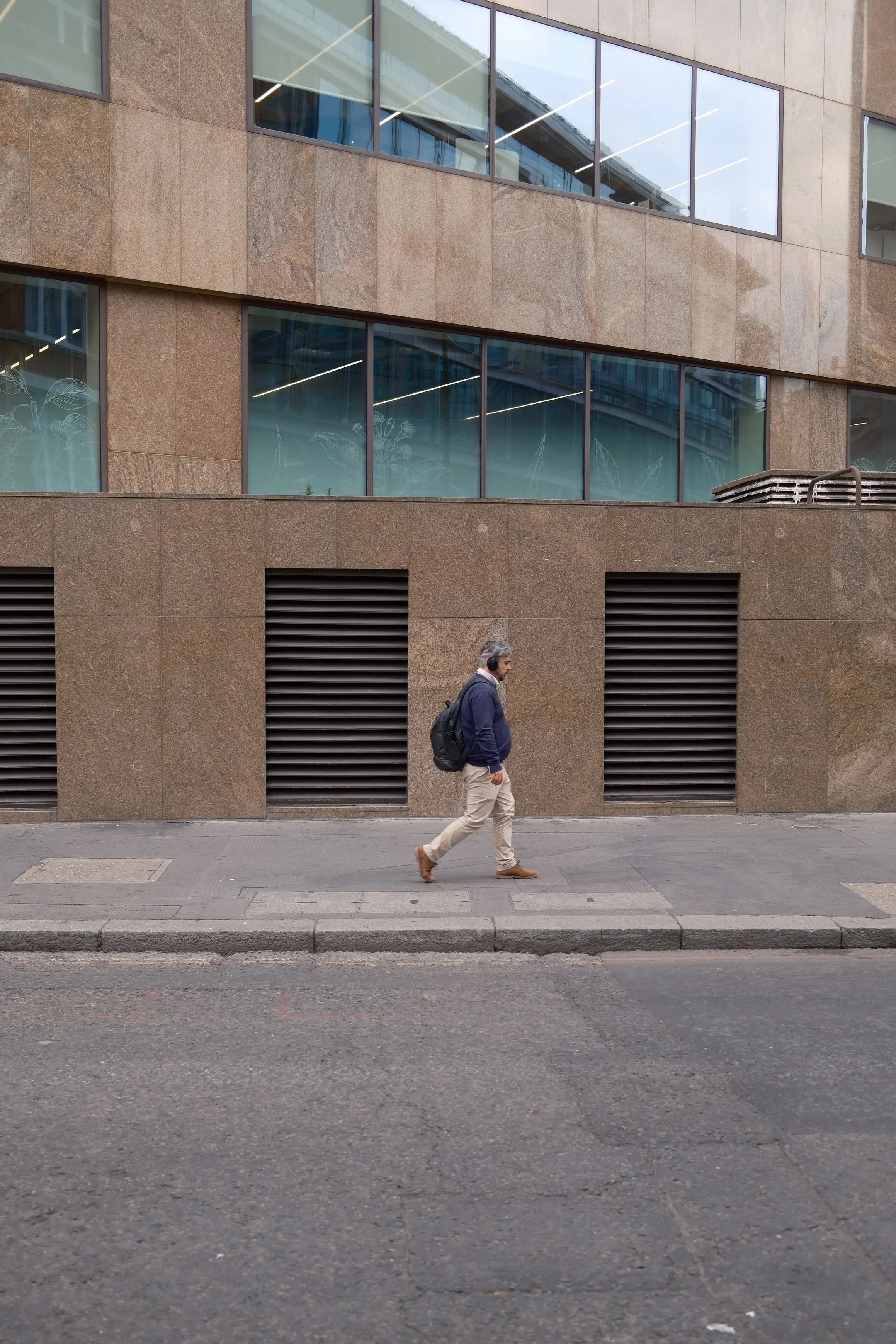 A man wearing a navy jacket, khaki pants, brown shoes, and headphones is walking on the sidewalk in front of a modern building with large windows and brown stone exterior.
