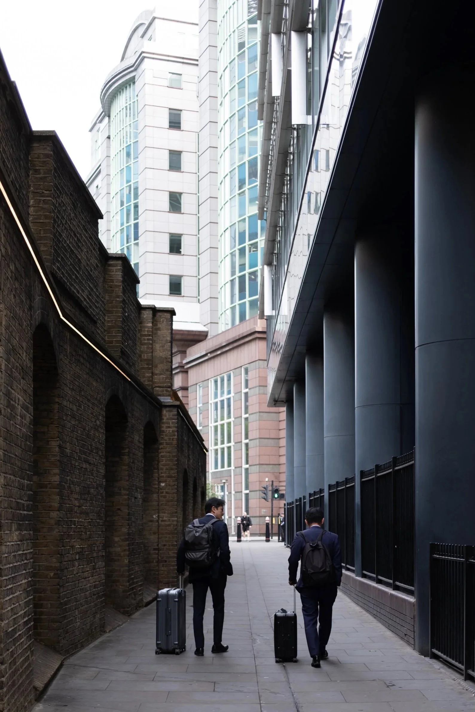 Two men in suits and backpacks walking with rolling suitcases down a city sidewalk. On the left is a brick building, and on the right is a modern glass and metal structure. In the background, there is a city street with a traffic light.