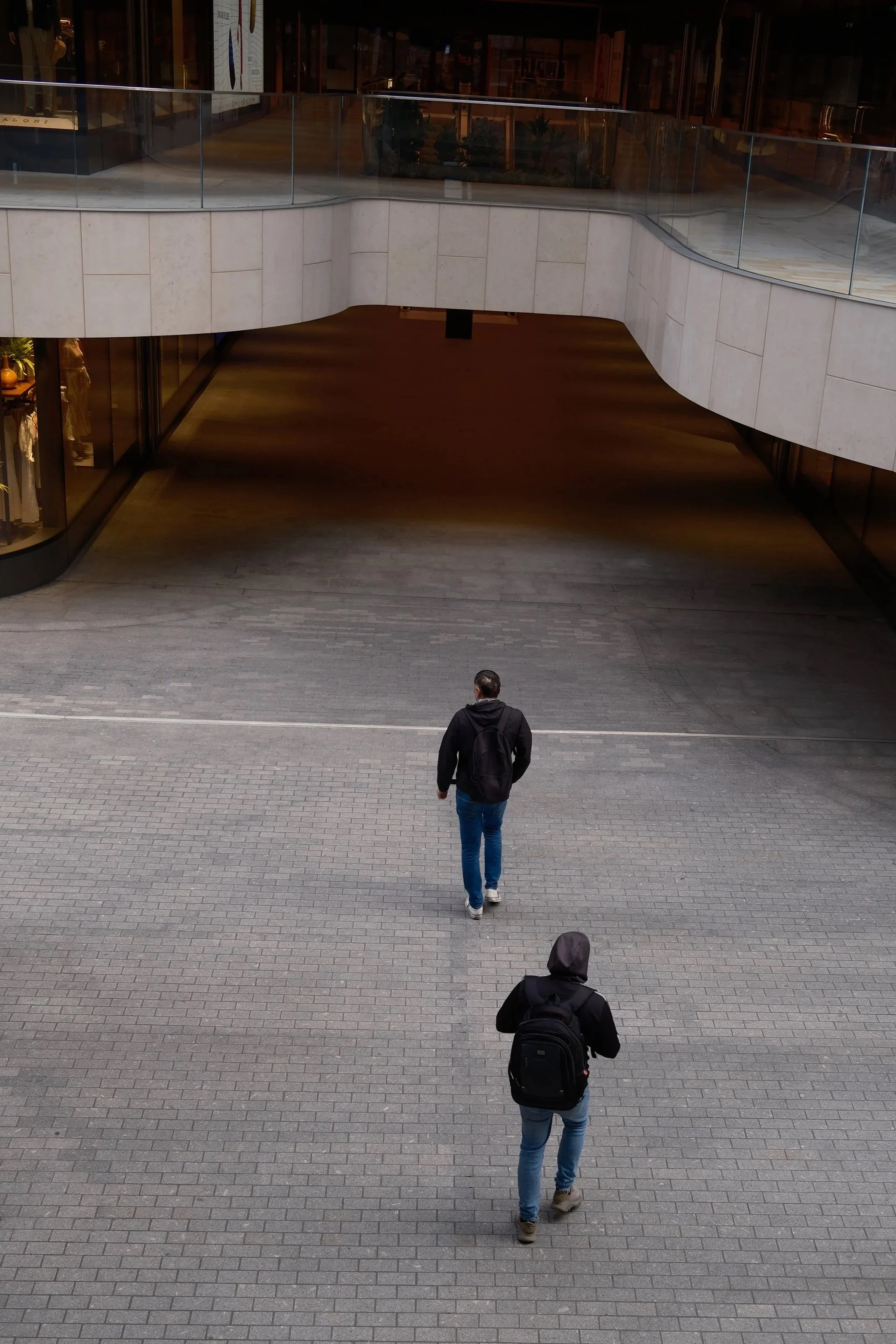 Three people walking away from the camera on a paved street. They are in front of a modern building with a curved, stone facade and glass railings.
