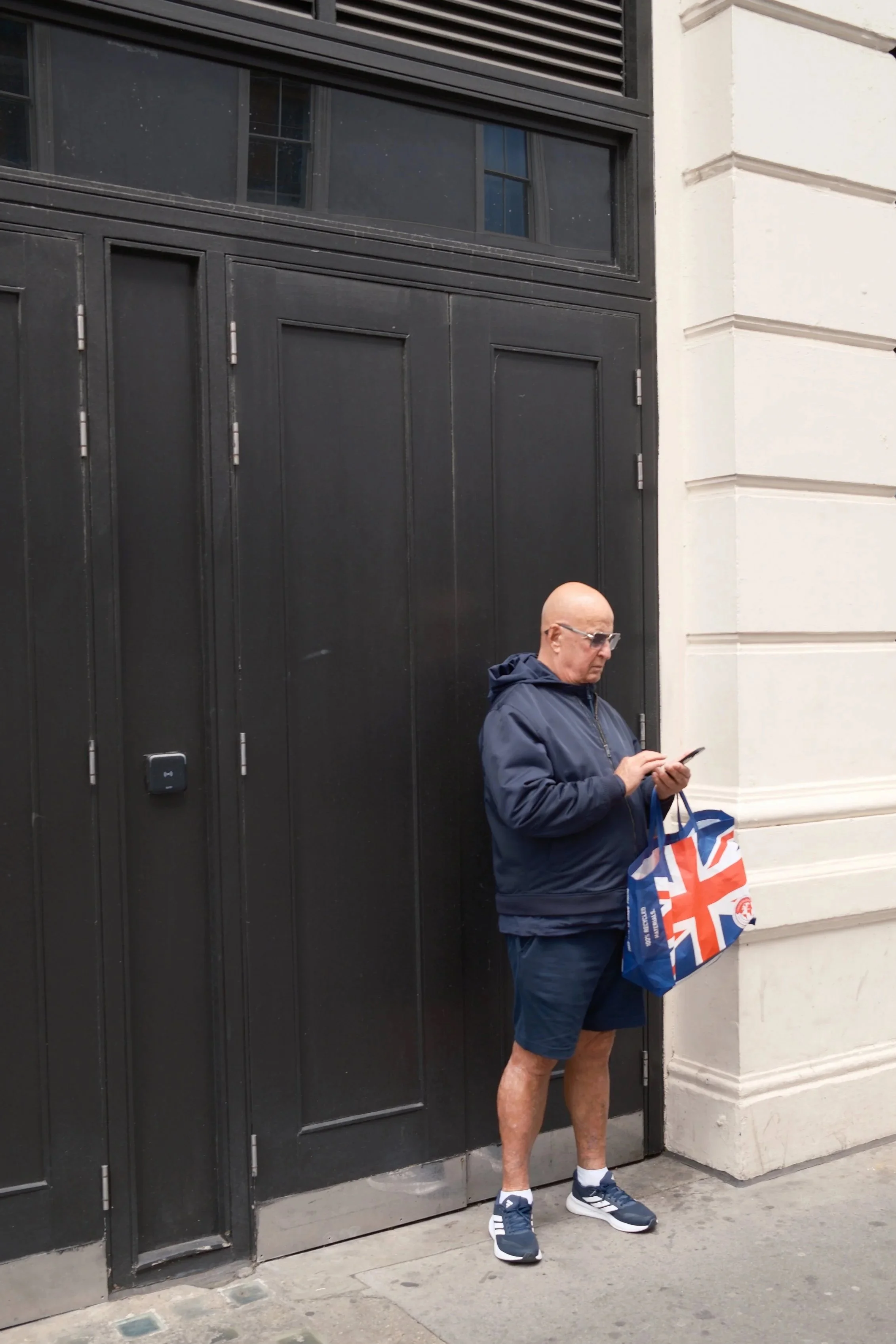 A man in a navy jacket, shorts, and running shoes is standing outside a black door, holding a blue shopping bag with the Union Jack design and looking at his phone.
