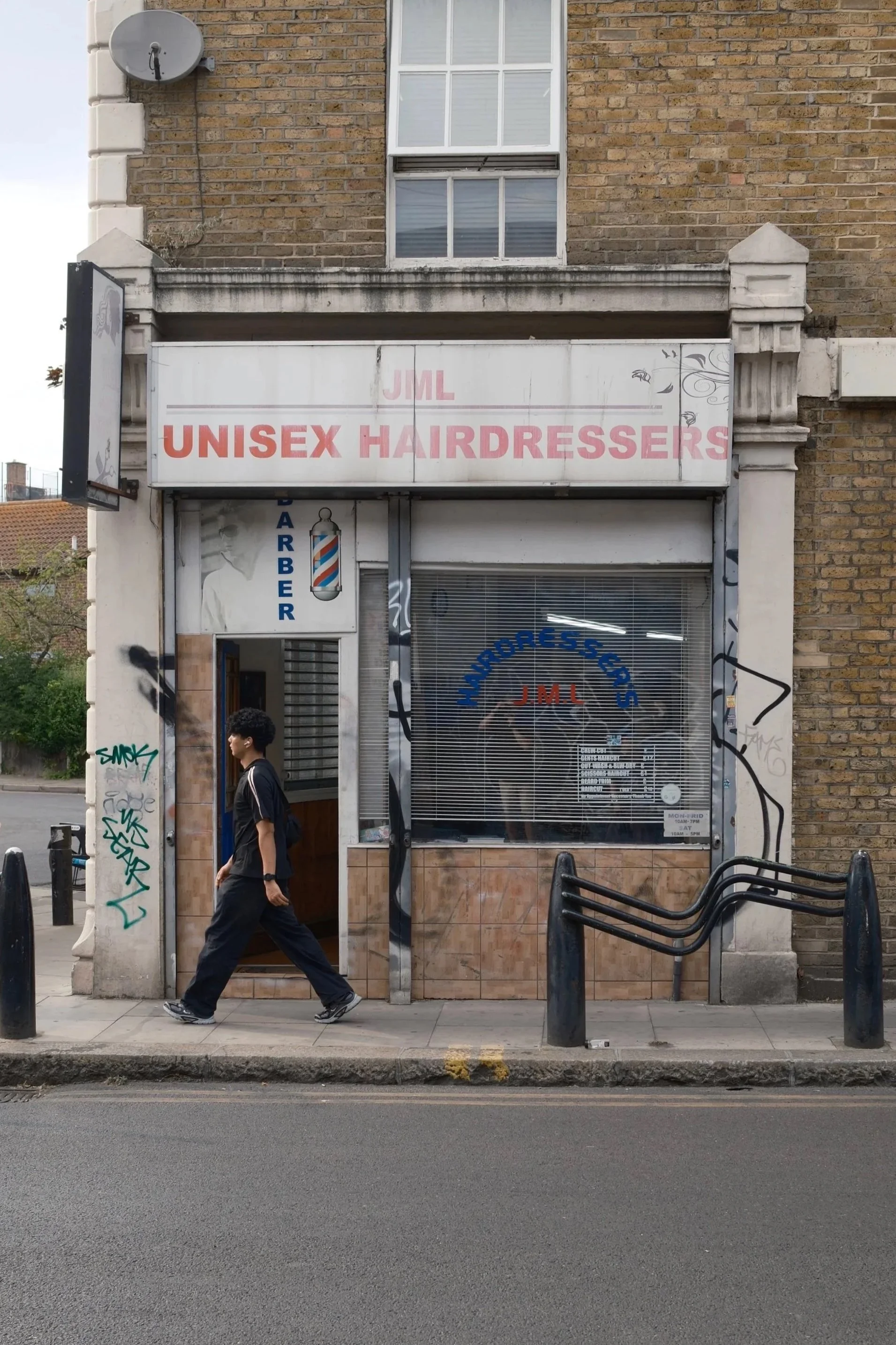 Street view of a storefront with a sign that reads 'JML UNISEX HAIRDRESSERS'. A man is walking past the shop on the sidewalk, which has black railings and concrete pavement. The building has a brick exterior with graffiti on the column and a window a