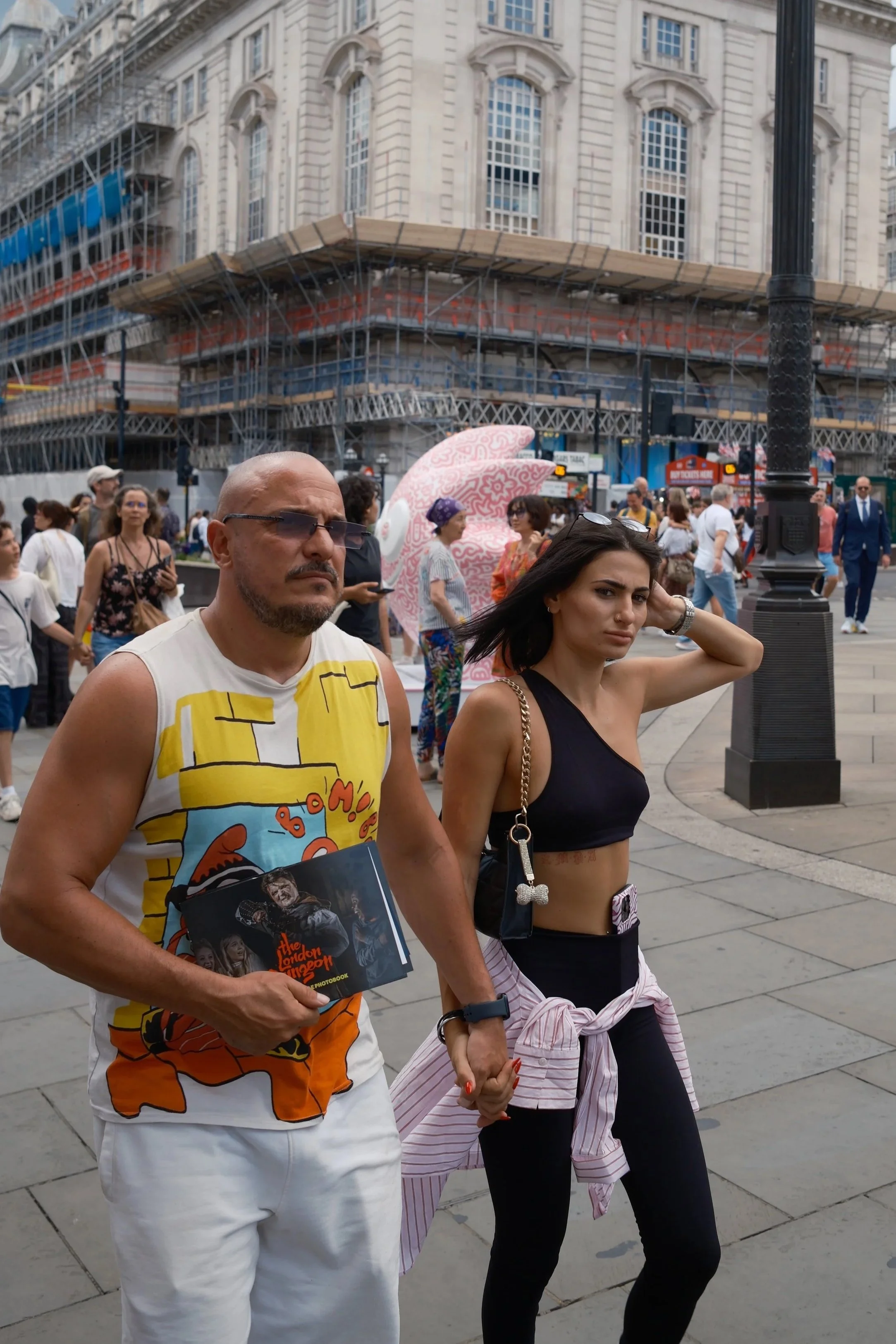 A man and a woman holding hands walking in a busy city street with a building under construction in the background, various pedestrians around, and a large pink and white sculpture. The man is wearing glasses, a colorful graphic sleeveless shirt, and