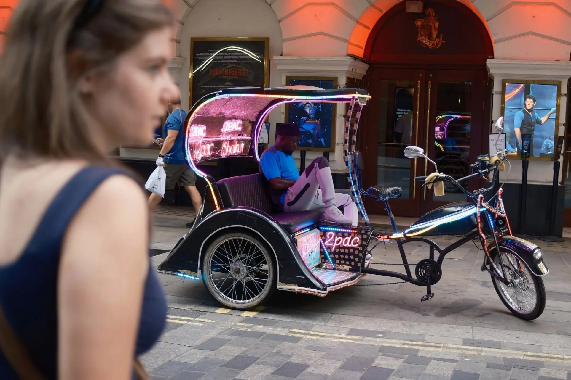 A colorful pedicab with neon lights and a sign that says "2Pac" is parked on a city street. A person is seated inside the pedicab, looking down at something, while a woman with brown hair, wearing a blue top, is walking past in the foreground. In the