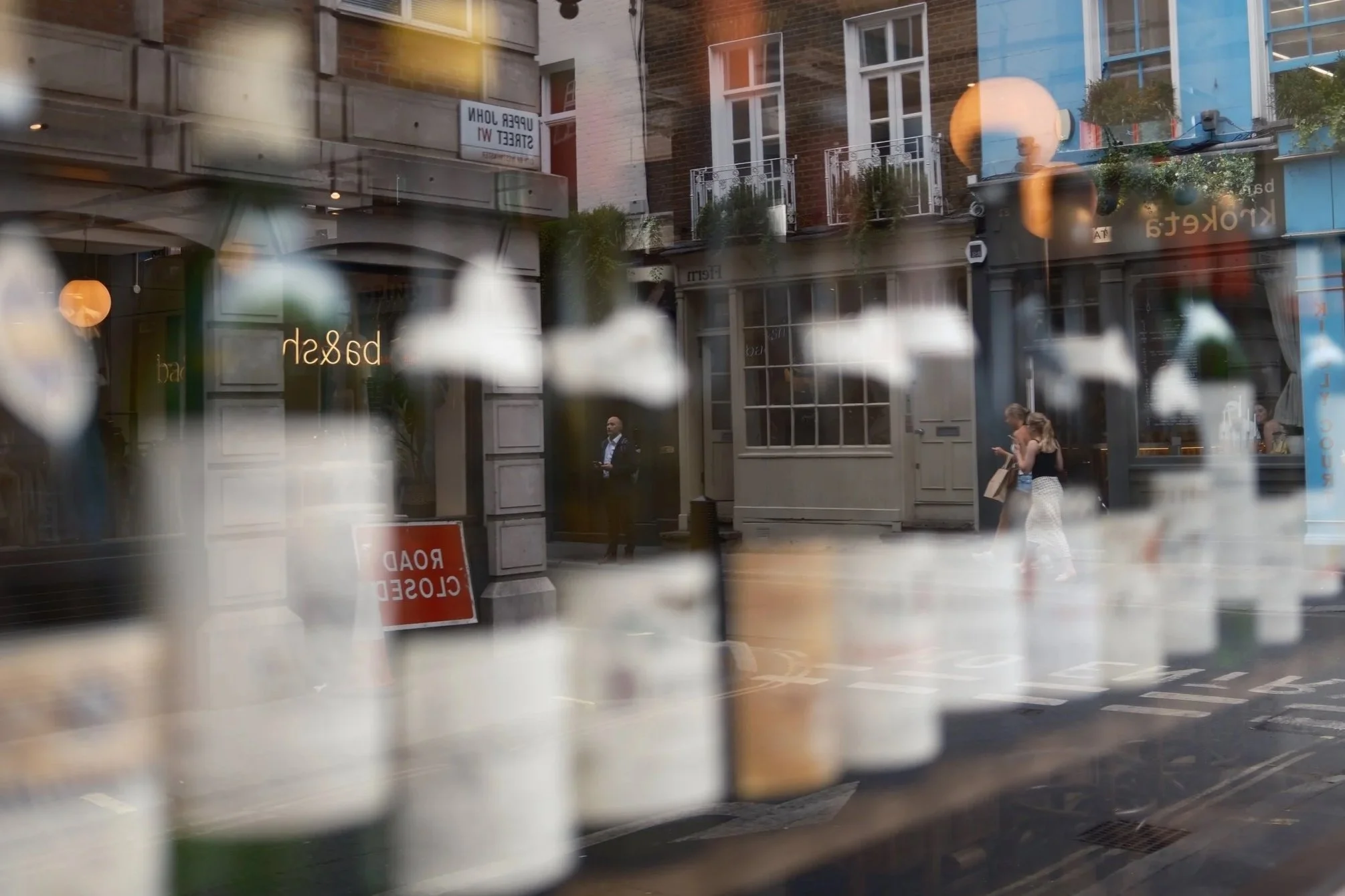 Reflection of street scene with people walking, buildings with window balconies, and a shop front with glass signage, seen through a window with white floral patterns.