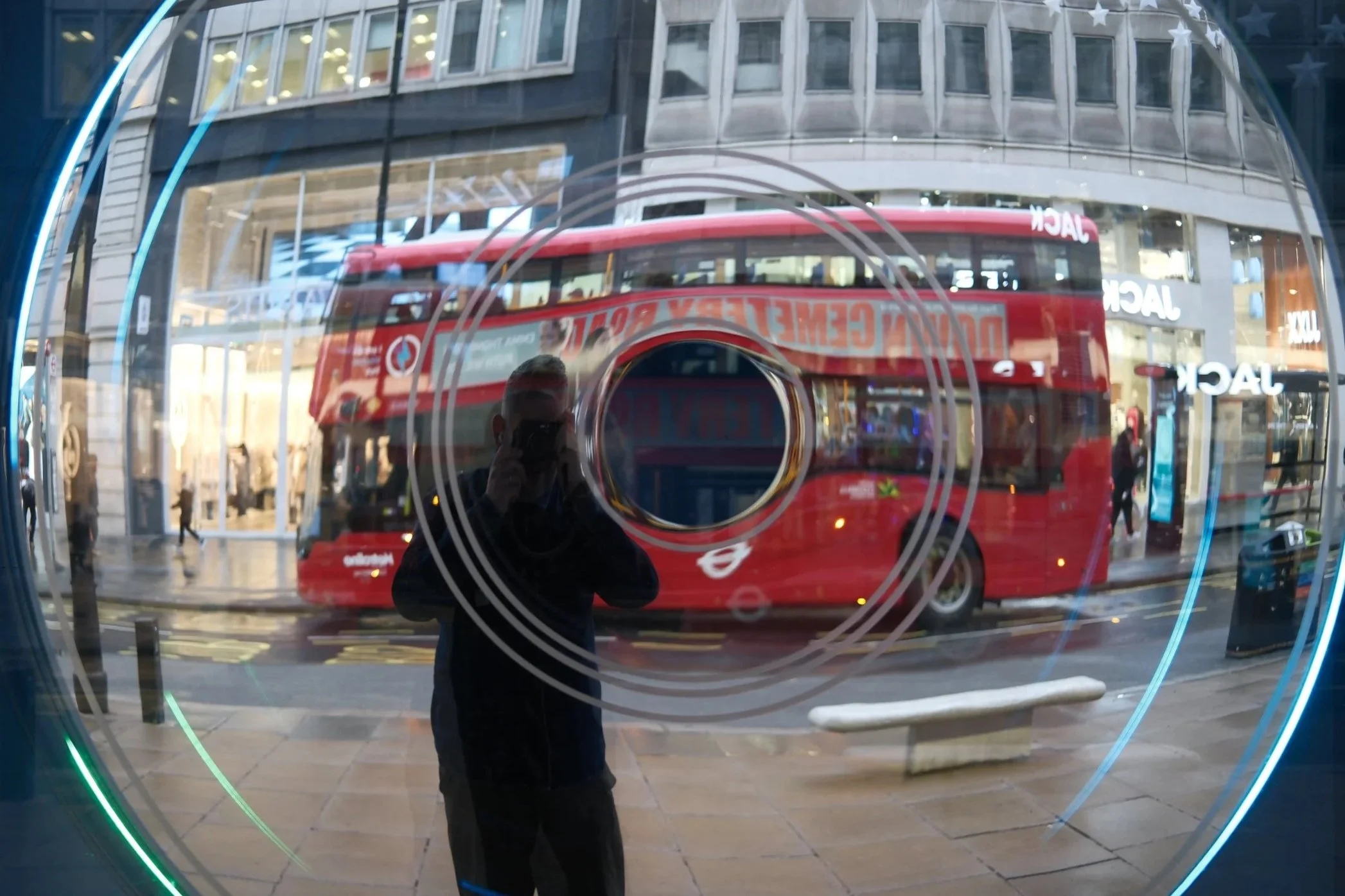 Reflection of a person taking a photo of a city street scene in a circular, glass window with a green neon light in the foreground. A red double-decker bus is visible through the glass, along with buildings and pedestrians.