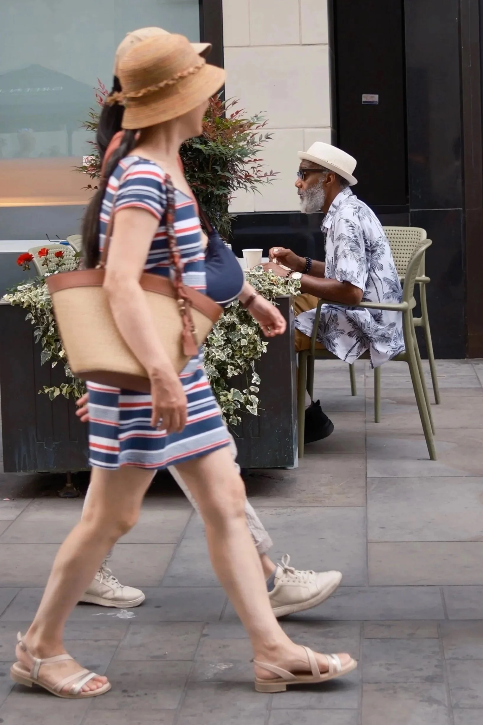 People walking on the street, one woman in a striped dress with a hat and another man sitting at a table, wearing a white hat and sunglasses.