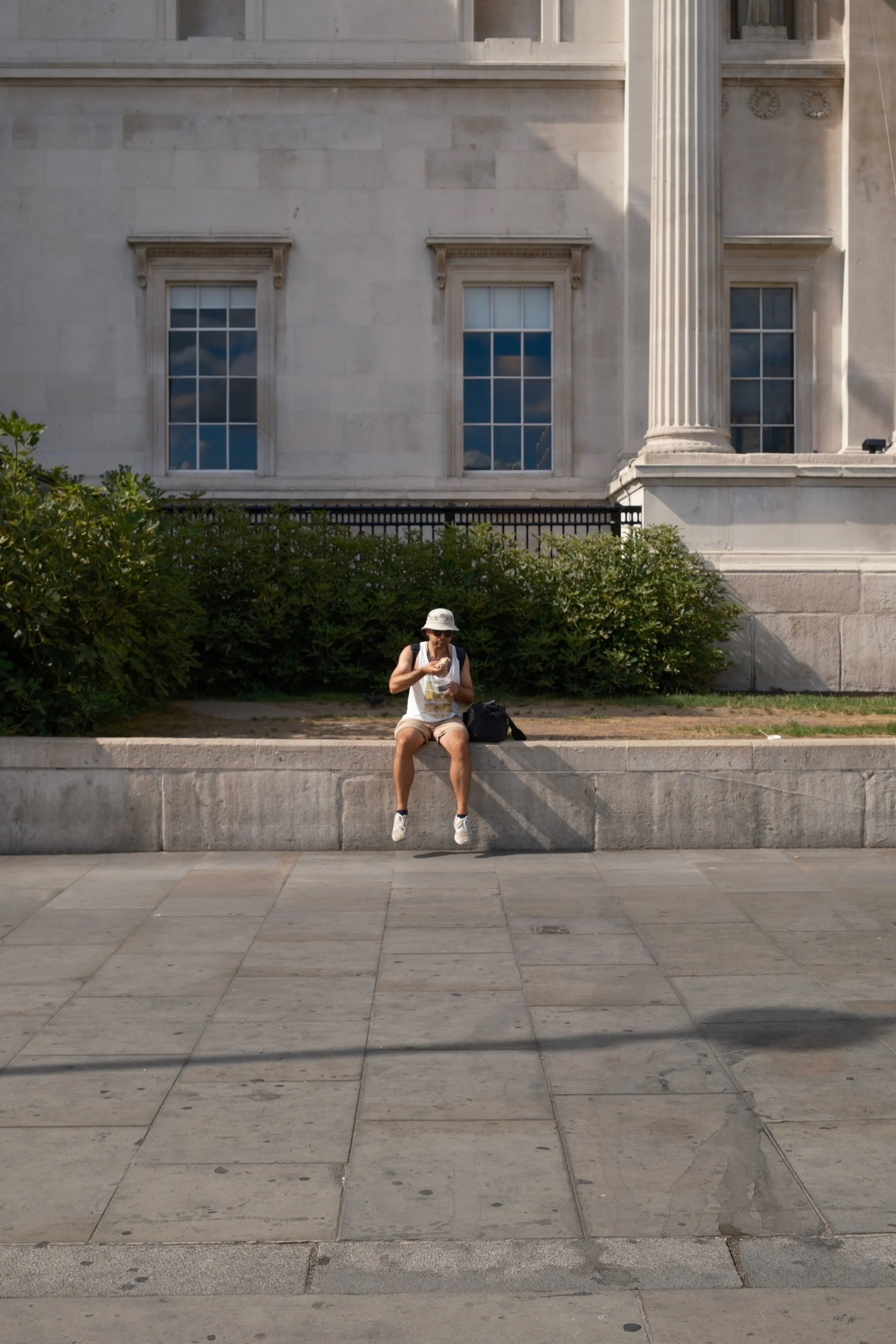 A woman with a white hat and white sleeveless top sitting on a concrete ledge, eating, with a building with large windows and columns behind her, and green bushes in front.